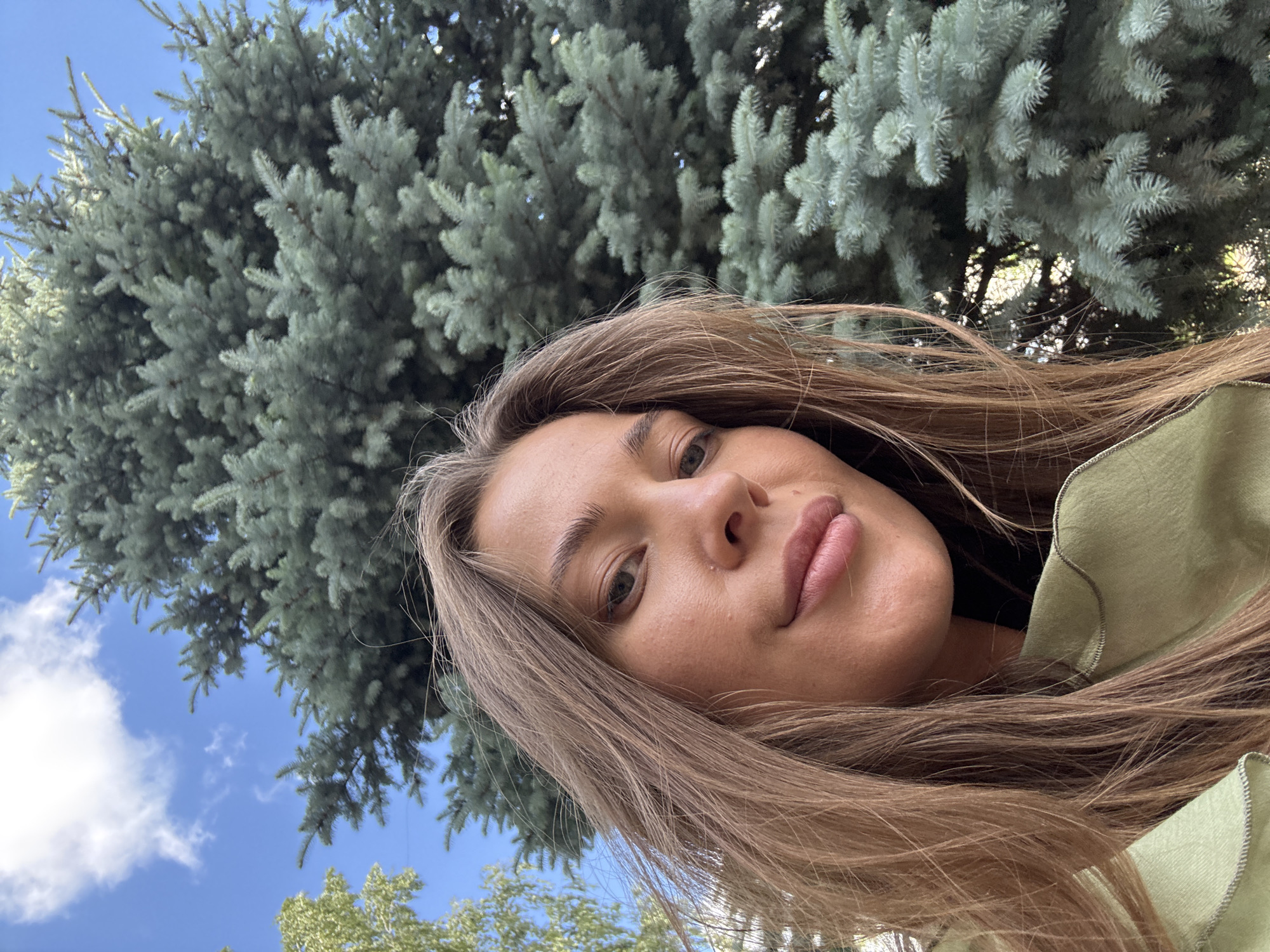 A woman's face is captured in a close-up, selfie-style shot, with her head tilted slightly to her right. She has long, light brown hair that falls around her face and shoulders. Her skin appears smooth with a light tan. She is wearing a light green top with ruffled edges. The background is dominated by a large, blue spruce tree with its needles clearly visible, and a patch of blue sky with a few white clouds. The image appears to have been taken outdoors on a sunny day. The location is identified as Chernushka, Russia, suggesting an outdoor setting within the city.