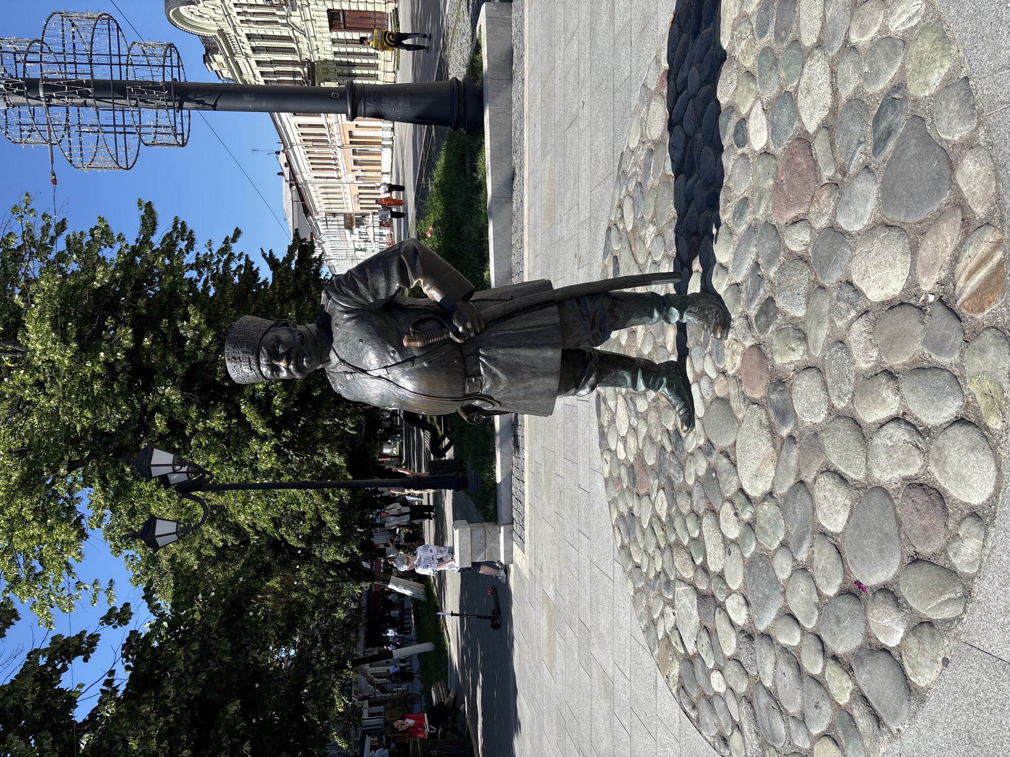 In the city of Oktyabrskoye, Russia, a bronze statue of a man in historical military attire stands prominently in a public square. The statue is positioned on a circular mosaic of smooth, grey river stones. The man is depicted wearing a tall, cylindrical hat with an inscription, a military tunic with buttons down the front, a belt with a buckle, trousers, and boots. He holds a sword by his side with his right hand. Behind the statue, a paved walkway is visible, lined with trees and street lamps. Further in the background, several people are walking, and buildings with traditional architecture are situated along the street. The scene is illuminated by natural sunlight, casting shadows on the ground.