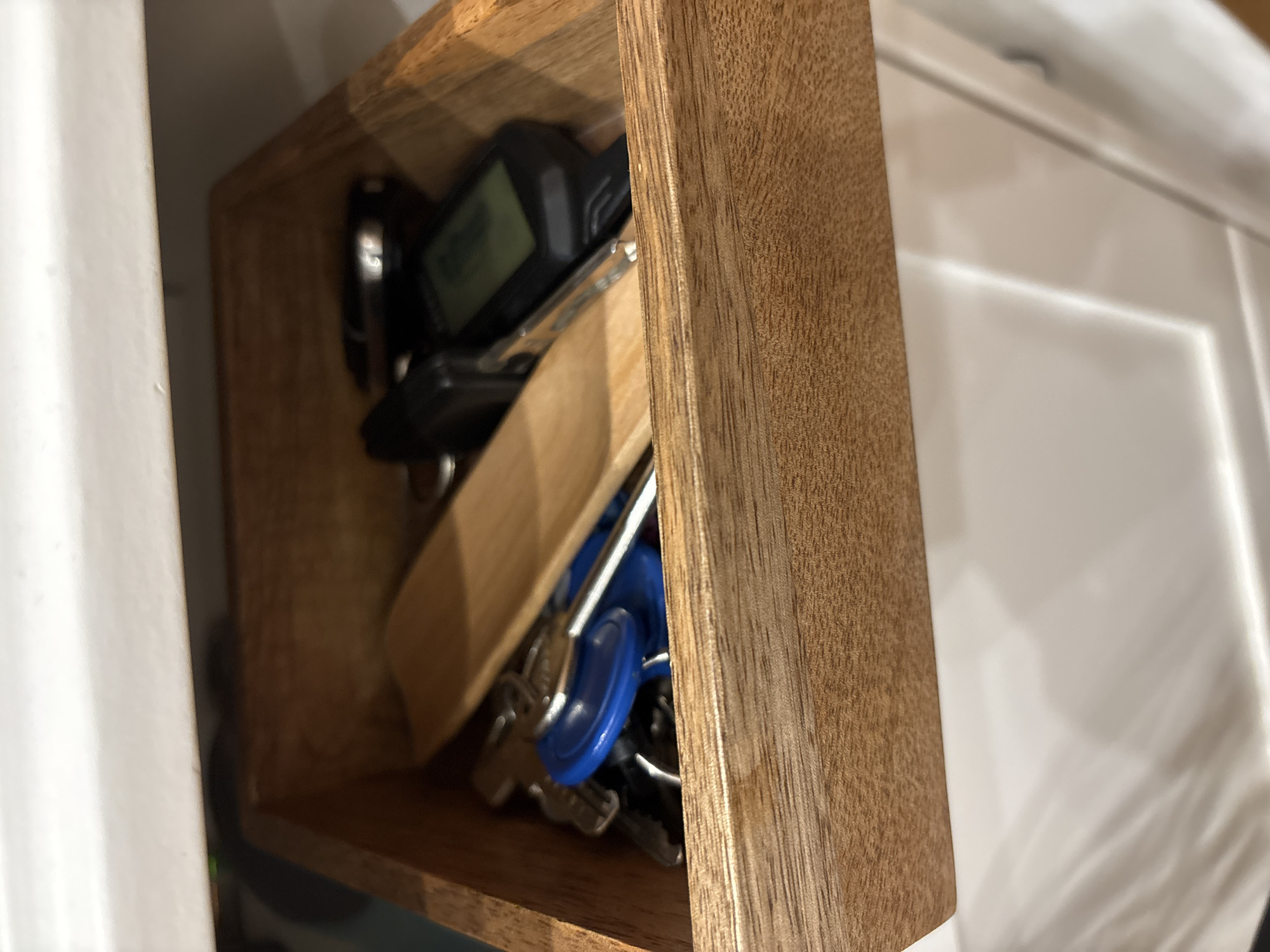 A close-up, slightly angled shot captures a collection of keys stored within a rectangular wooden box. The box, crafted from light-colored wood with visible grain, appears to be an organizer or key holder in an indoor setting.
Inside the box, various keys are jumbled together. These include several standard metal keys, a distinct blue plastic key cover or fob, and a modern black car remote or key fob. The car remote has a small, dark digital screen, but any text or numbers on it are too blurry to decipher. A smaller wooden element or divider is also visible within the box, contributing to its internal structure.
The background to the right of the box features a reflective, light-colored surface, possibly a cabinet or appliance, showing subtle reflections. To the left, a plain white wall or cabinet edge provides a clean, contrasting backdrop. The lighting indicates an indoor environment, likely illuminated by artificial light. No activity is taking place; the image simply presents a static arrangement of stored items. There are no identifiable visual cues within the image to suggest a specific geographical location.