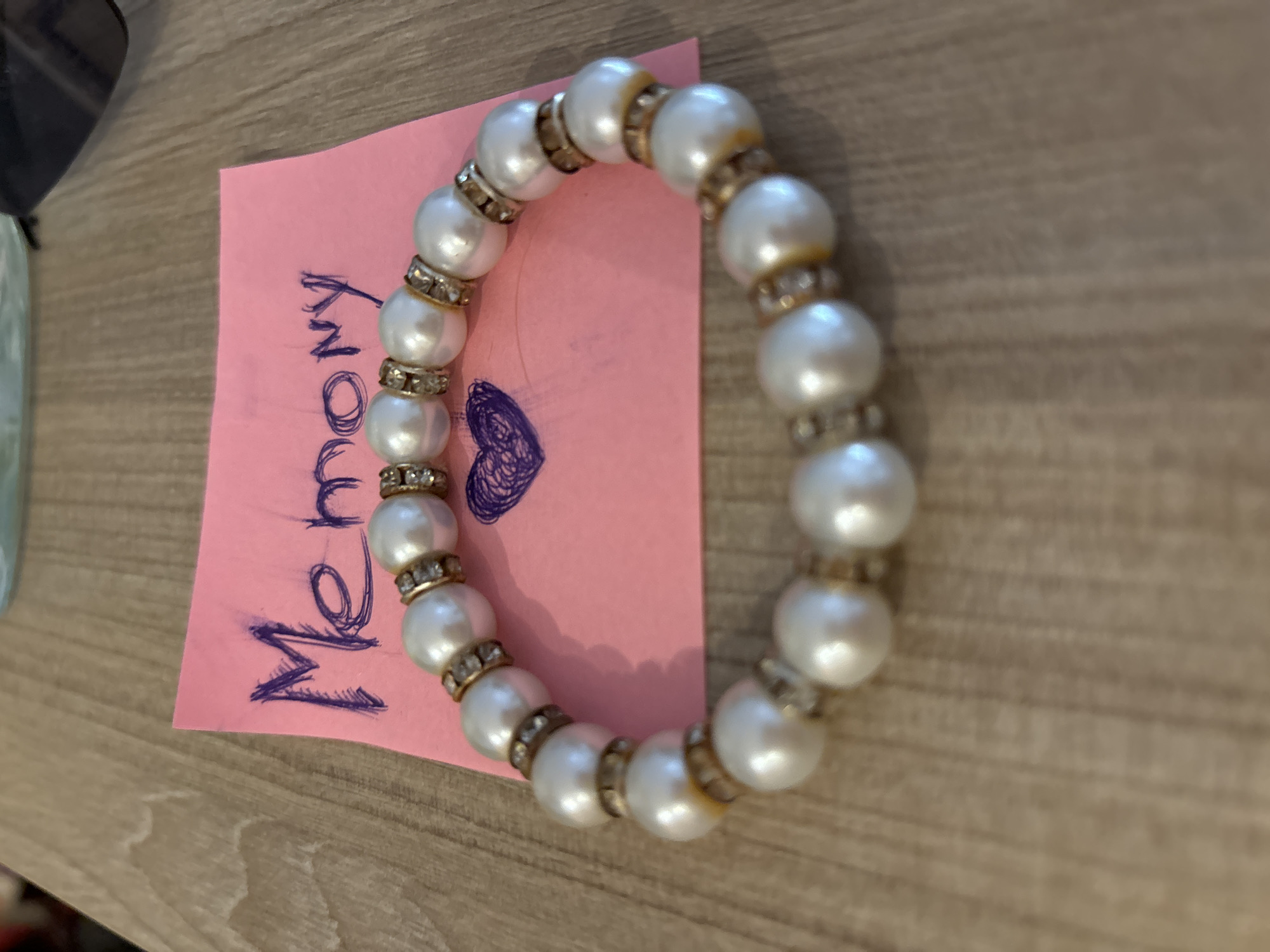 A close-up, eye-level shot shows a pearl bracelet resting on a pink sticky note with the word "Memory" handwritten in purple marker. A small, hand-drawn heart is also present on the note. The background consists of a light brown wooden surface. The scene is indoors, and there are no people or animals visible. The lighting appears to be natural, casting soft shadows. The image is sharp and in focus on the bracelet and sticky note.