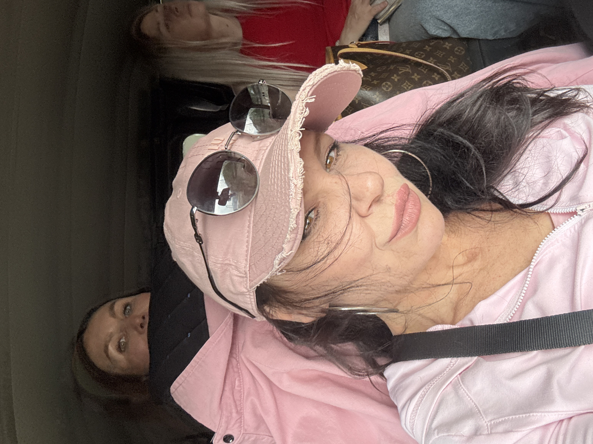 Three women are seated in a vehicle. The woman in the foreground, wearing a pink baseball cap and sunglasses, is looking towards the camera. She has dark hair and is wearing a pink jacket. Behind her, to the left, another woman with brown hair is looking upwards, with only her eyes and forehead visible above a black collar. To the right, a woman with blonde hair wearing a red shirt is looking forward. A Louis Vuitton patterned bag is visible between the foreground woman and the woman on the right. The scene appears to be captured during daylight, with reflections of the outdoors visible in the sunglasses. There is no information in the image to suggest a specific location in Каменномостское сельское поселение, Russia.
