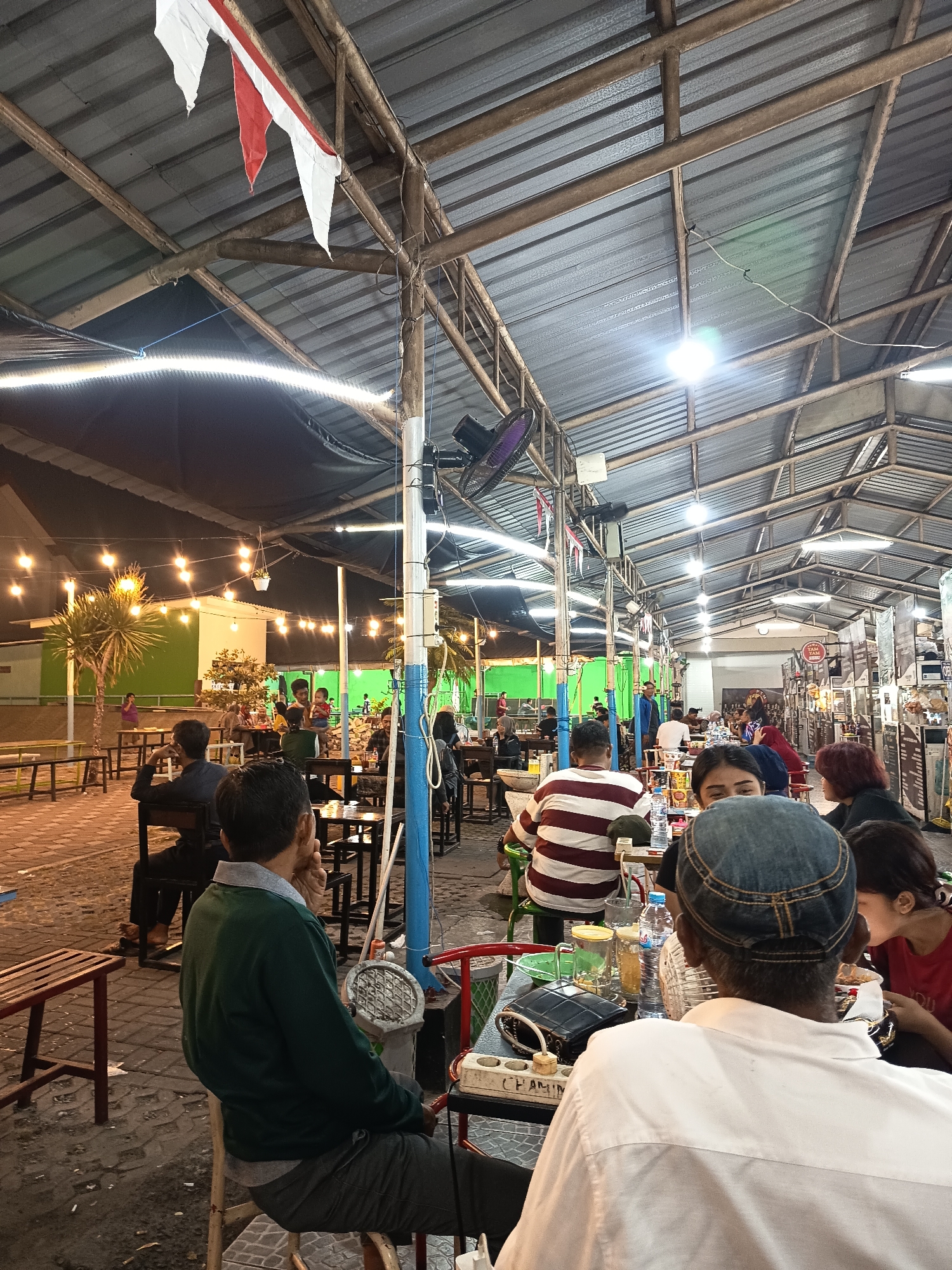 The image depicts an outdoor food court or restaurant in Surabaya, Indonesia, at night. The scene is covered by a corrugated metal roof supported by metal and bamboo structures. String lights and industrial lighting illuminate the area. Several people are seated at tables, appearing to be dining or socializing. In the foreground, a man wearing a white shirt and a denim cap is facing away from the camera, seated at a table with food and drinks. To his left, a person with dark hair is wearing a red shirt, and to his right, another person is visible from behind. The environment shows a brick-paved ground with scattered debris. In the background, more tables, people, and palm trees are visible, along with green-painted walls and buildings, suggesting an urban setting. A fan is mounted on a pole to the left, and triangular red and white flags are strung from the ceiling.