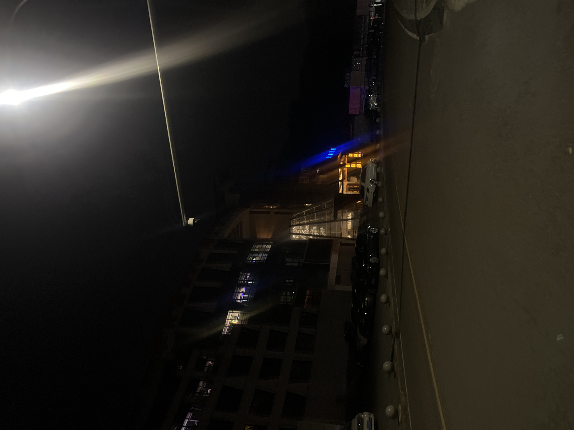 The image displays an urban nighttime scene in Jiulongpo District, China, featuring a paved road, parked vehicles, and a multi-story building. A bright street light positioned in the upper right corner casts a strong vertical light beam and illuminates the foreground.
On the left, a large, dark building, likely a commercial or hotel structure, features numerous windows; some display interior lighting, creating visible rectangles of light. The building exhibits modern architecture, including a prominent glass-fronted section or atrium towards the center, which is also internally lit with a warm, yellowish glow.
Along the edge of the road in the midground, a series of light-colored, spherical concrete bollards separate the roadway from a parking area. Multiple vehicles, predominantly dark-colored sedans and at least two white sedans, are parked in this area. The road itself is marked with a continuous yellow line.
Further to the right, an adjacent structure or section of the building emits a distinct blue light from its facade, contrasting with additional warm yellow/orange lighting near an entrance. A white security camera or similar device is mounted on an overhead pole or structure in the upper-middle frame. No people are visible within the scene.