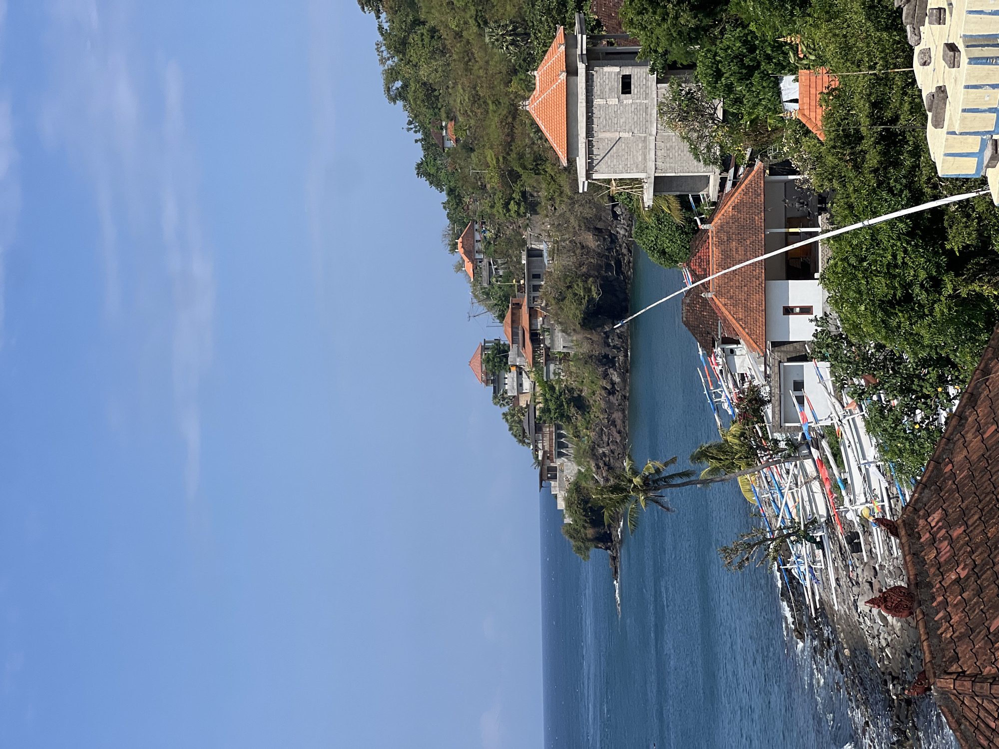 A bright, sunny day in Abang, Indonesia features a rocky coastline with numerous small, white boats lined up on the shore. The ocean is a deep blue, with gentle waves lapping against the shore. A lush green hillside rises from the coastline, dotted with several buildings, most featuring traditional orange-tiled roofs. Some of these structures appear to be residences, while others may be religious buildings. In the foreground, a prominent tiled roof with intricate decorative elements is visible. A tall, unfinished concrete structure with an orange roof stands out on the right side of the frame, indicating ongoing construction. The sky is a clear, vibrant blue with a few wispy white clouds. The scene conveys a sense of coastal life and development in a tropical setting.