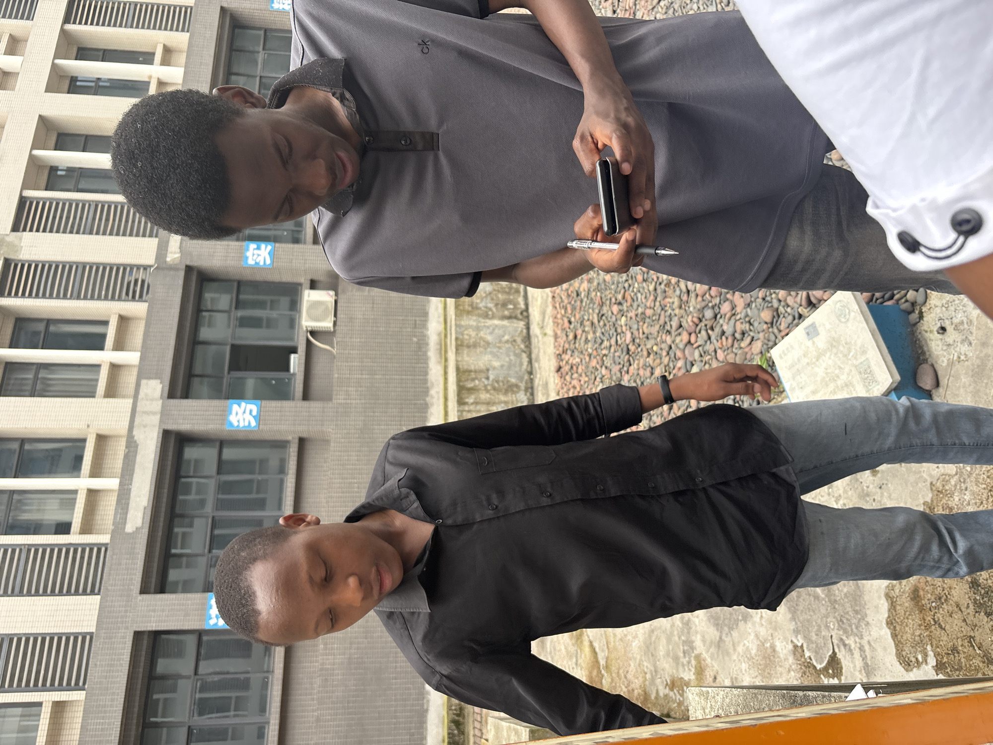 This image captures an outdoor scene featuring two young men and a partial view of a third person, set against a backdrop of modern multi-story buildings. The lighting indicates it is daytime.
The man in the foreground on the left wears a gray polo shirt with a "CK" (Calvin Klein) logo, dark jeans, and has short, dark curly hair. He is focused downwards, holding a small black electronic device (possibly a phone or vape pen) in his left hand and a pen in his right. Beside him, the man in the foreground on the right wears a black long-sleeved button-down shirt and light blue/gray jeans, with a dark wristband on his left arm. His eyes are closed or looking down, conveying a calm or reflective expression. A third person is partially visible on the far right, wearing a white long-sleeved shirt with black buttons.
The ground is a mix of reddish-brown gravel and concrete. In the background, several contemporary buildings with numerous windows and balconies/louvers rise. An air conditioning unit is visible on one building. Blue Chinese characters are repeatedly displayed on the building walls; these characters spell "教育" (jiàoyù), which translates to "education," strongly suggesting the location is an educational institution like a university or school in Jiangjin, China. A white book or document lies on the ground near the partial figure. The overall atmosphere appears casual and observational, with the men seemingly engaged in individual activities or moments of pause.
