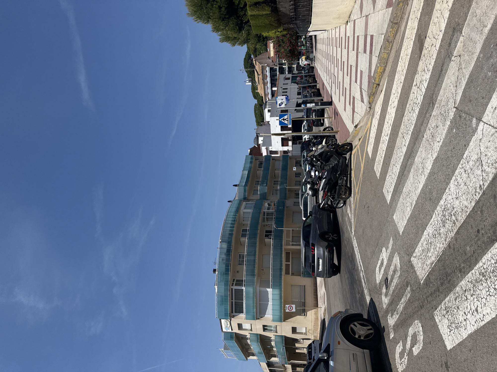 A street scene in Castell d'Aro, Platja d'Aro i s'Agaró, Spain, under a clear blue sky with scattered wispy clouds. In the foreground, a crosswalk is marked with white stripes, and the word "STOP" is painted on the asphalt. A silver car is parked to the left, partially visible. Behind the crosswalk, a gray SUV is driving, and several motorcycles are parked on the right side of the street. A building with a curved facade and turquoise balconies dominates the left side of the image. Several other buildings, some with white walls and tiled roofs, are visible further down the street. A sidewalk with a patterned tile design runs along the right side of the street, bordered by a hedge. Trees are visible on the right, and a speed limit sign of 30 km/h is posted on the left side of the street.