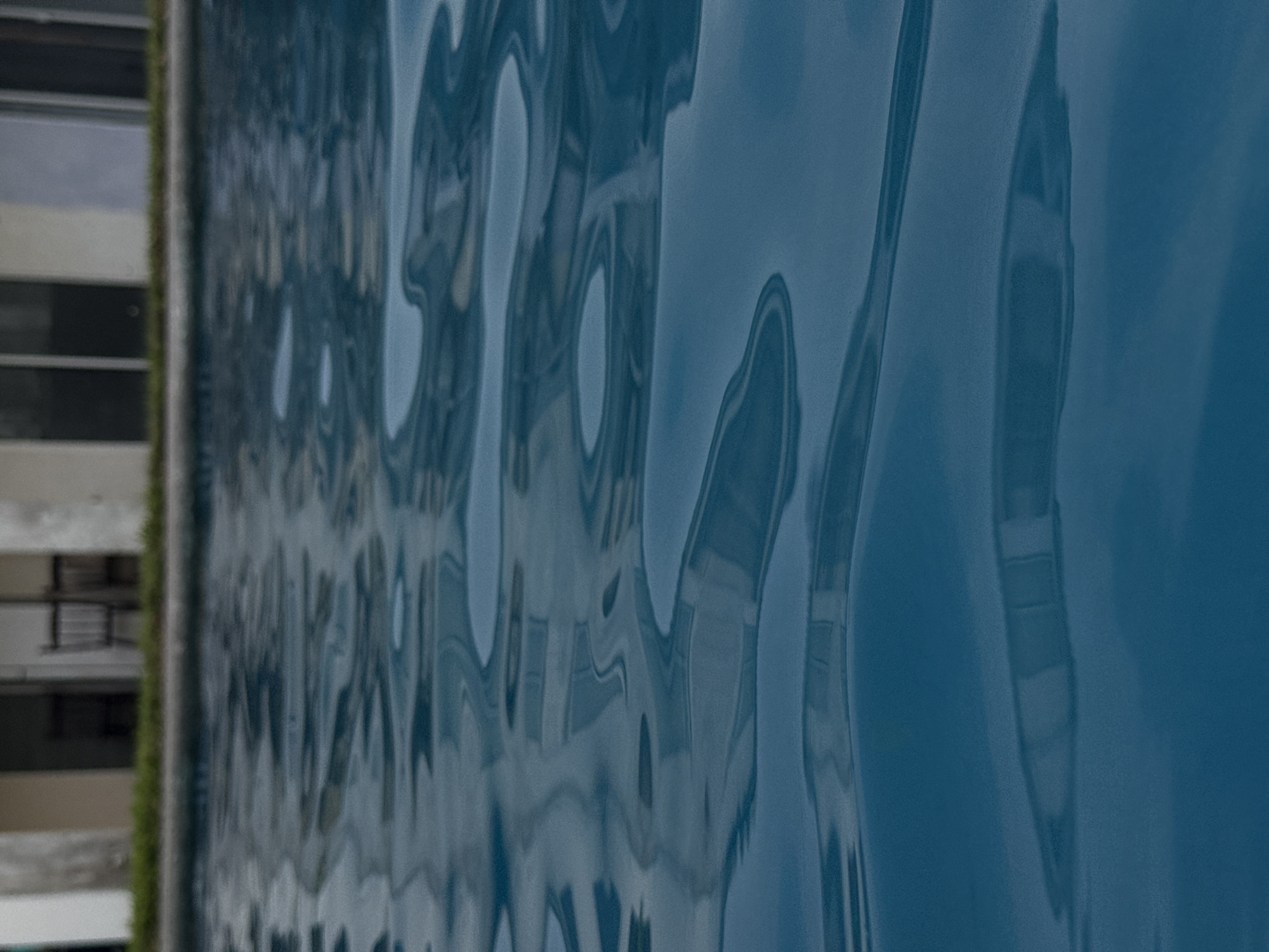 A long, vertical shot shows the reflections of a building, windows, and sky on the surface of a pool in Veracruz City, Mexico. On the left of the shot, a tall building is seen with windows lined up. On the surface of the water, there is a ripple effect. The color of the water is a dark blue-gray, and the reflections shimmer on the surface of the water. The left border of the pool is green. The angle of the pool causes the building to appear at a tilted angle.