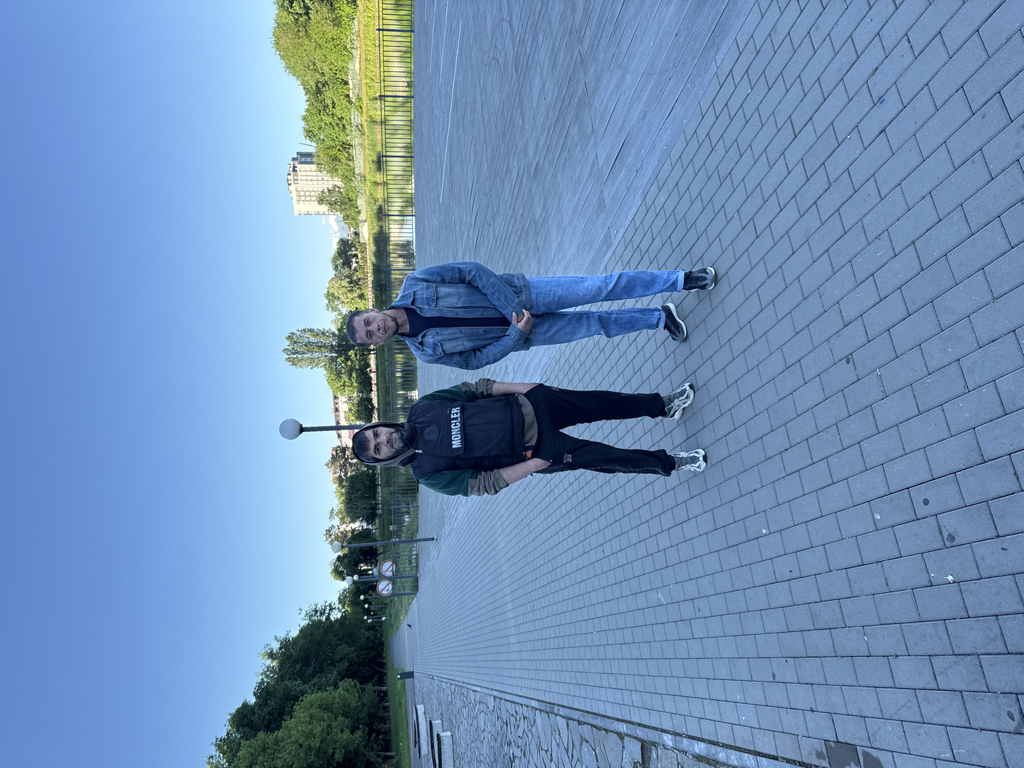 Two men stand on a gray-bricked walkway with trees and a building visible in the background, under a clear blue sky. One man wears a denim jacket and jeans, while the other is dressed in a black vest over a green sweatshirt and black pants. The location appears to be a park or public area in an urban setting.