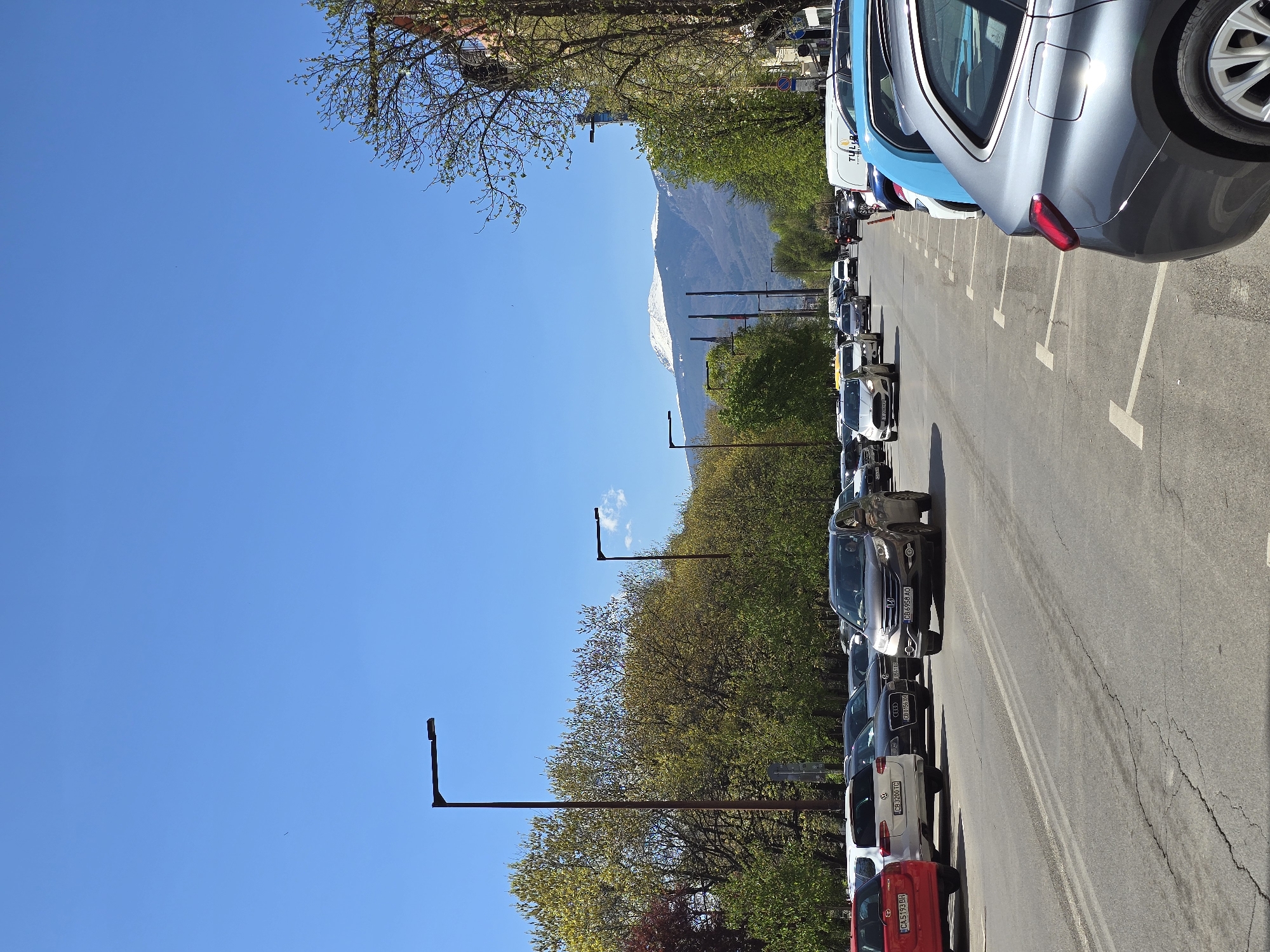 The image depicts a parking lot in Sofia, Bulgaria, filled with parked cars. A mountain range with snow-capped peaks is visible in the background. Trees line the parking area. The sky is clear and blue. Street lamps stand along the parking lot.
