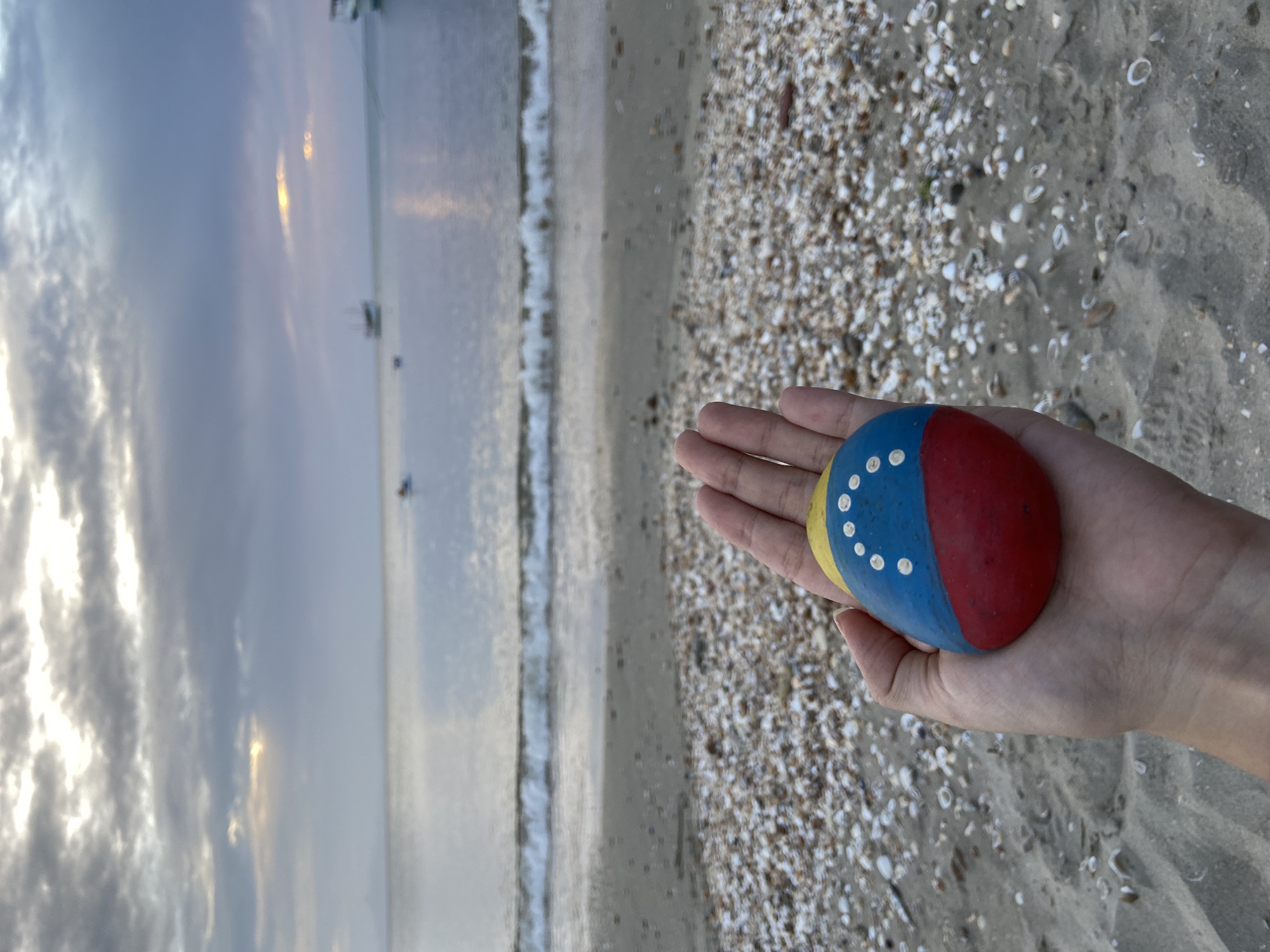 A hand holds a painted stone, designed to resemble the Venezuelan flag, against a backdrop of a beach and the sea. The stone, with its distinct yellow, blue, and red stripes and white stars, is prominently displayed in the foreground. The beach is covered with small shells and sand, with gentle waves lapping at the shore. In the distance, several boats are visible on the calm water under a cloudy sky. The lighting suggests it may be dawn or dusk, casting a soft glow over the scene. The location is identified as Juan Griego, Venezuela, a coastal town known for its picturesque beaches and natural beauty.