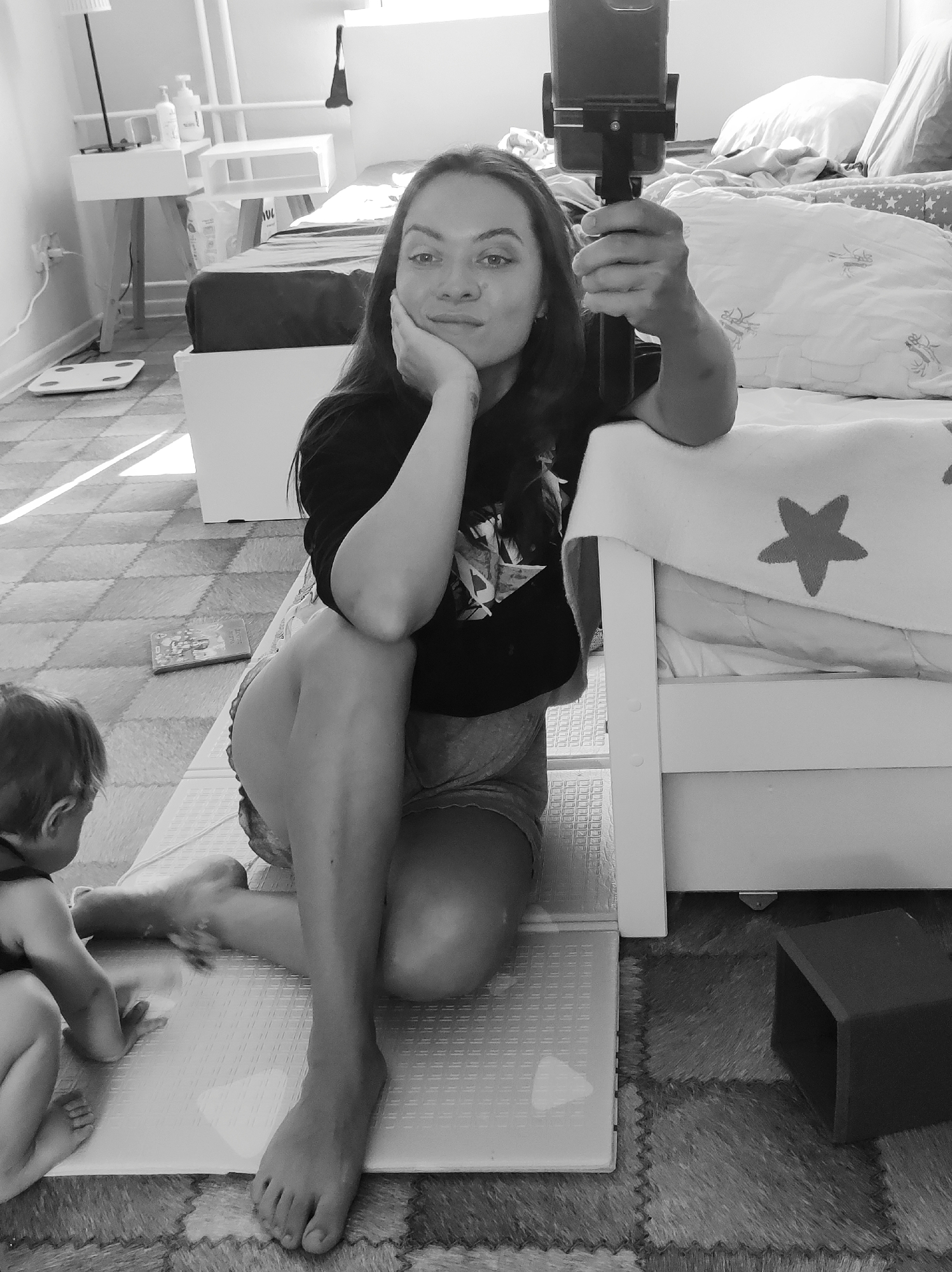 A young adult female is sitting on the floor with a child, holding a phone and tripod, presumably taking a selfie or video. She is wearing a dark t-shirt and shorts, and is resting her head on her hand. The child is seated on the floor. Behind the woman is a bed with a white frame. The room also contains a bedside table, a scale, and a toy. The location is indicated as Chaikovsky, Russia.