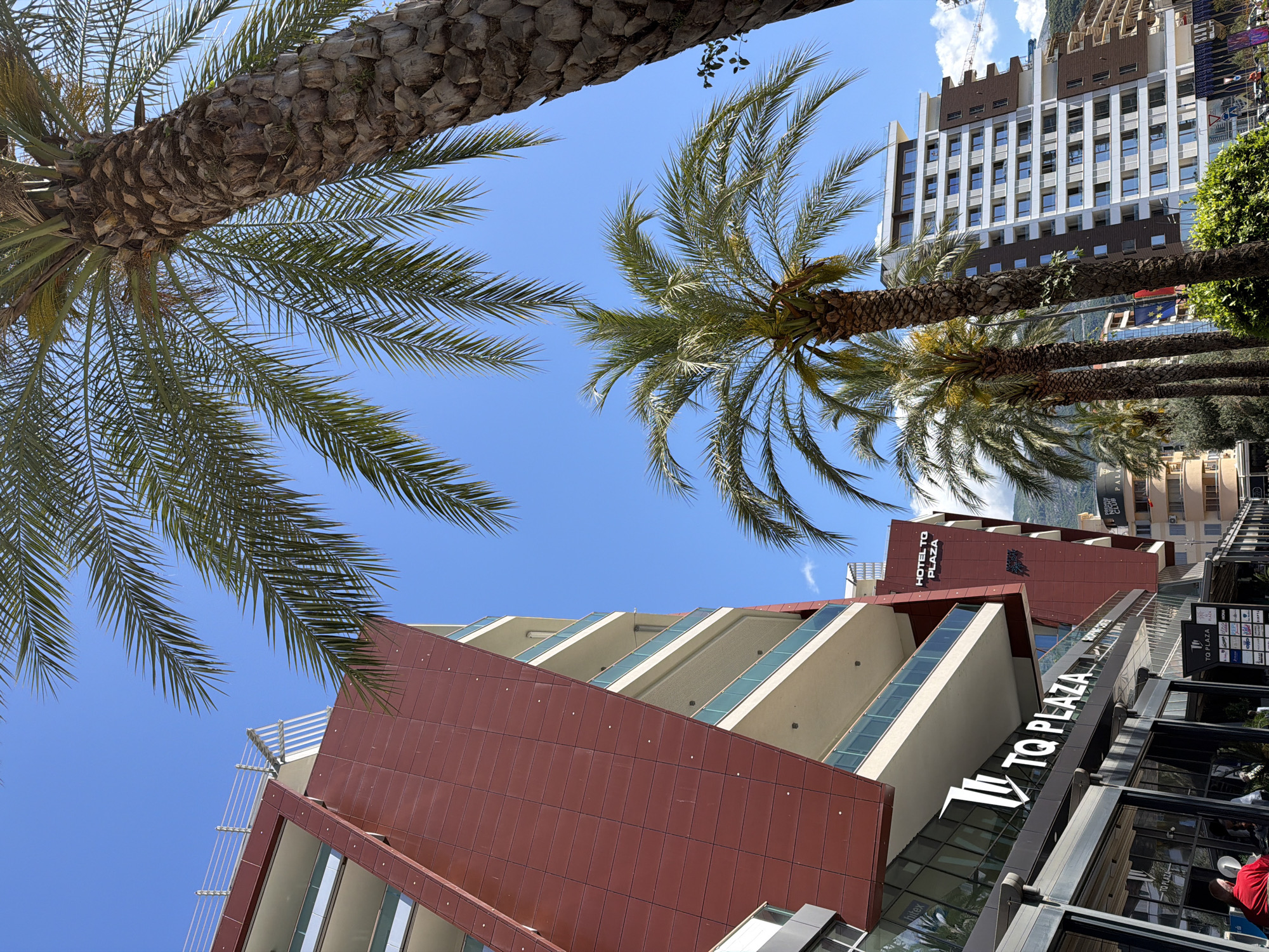 The image, taken from a low angle, showcases a modern hotel named "TQ Plaza" in Budva, Montenegro. The hotel has a geometric design with reddish-brown paneling and glazed sections. Part of a taller building is visible in the background. Palm trees frame the composition, creating a tropical atmosphere. The sky is blue with a few clouds. The image suggests a sunny day in an urban setting known for its coastal tourism.