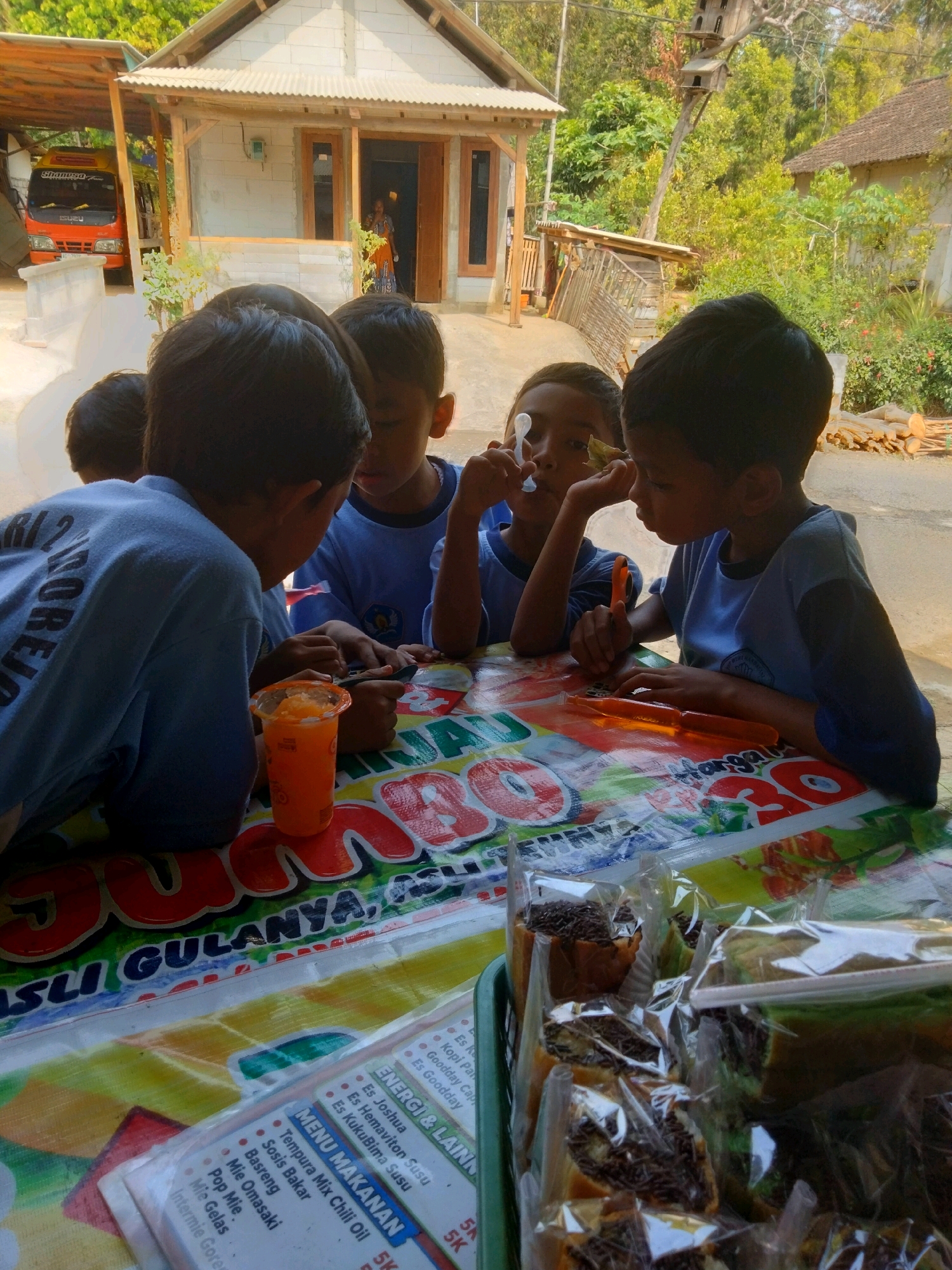 The image depicts a group of four young boys gathered around a table in what appears to be the city of Sidorejo, Indonesia. The table is covered with a colorful, patterned plastic tablecloth. In the foreground, on the table, are several plastic-wrapped food items, including what looks like bread with chocolate sprinkles and a green cake. To the left, an orange beverage in a plastic cup is visible.
The boys are all wearing blue t-shirts. Three of the boys are facing the table, engaged with something on it, possibly food or game pieces. One boy in the foreground on the left is leaning on the table, his back to the camera, with the text "ORI" visible on his shirt. Another boy in the center is looking down at his hands on the table. To his right, a third boy is also looking down at the table. The fourth boy on the right is turned slightly away from the table, with his face partially visible.
In the background, a simple house with a corrugated metal roof and white brick walls is visible. A red and yellow Isuzu truck is parked to the left of the house, partially under a lean-to structure. The surrounding environment is lush and green, with trees and vegetation, suggesting a rural or semi-rural setting within Sidorejo. A person in a patterned orange and blue garment can be seen through the open doorway of the house. The overall scene conveys a casual, everyday moment of children gathering around a table outdoors.