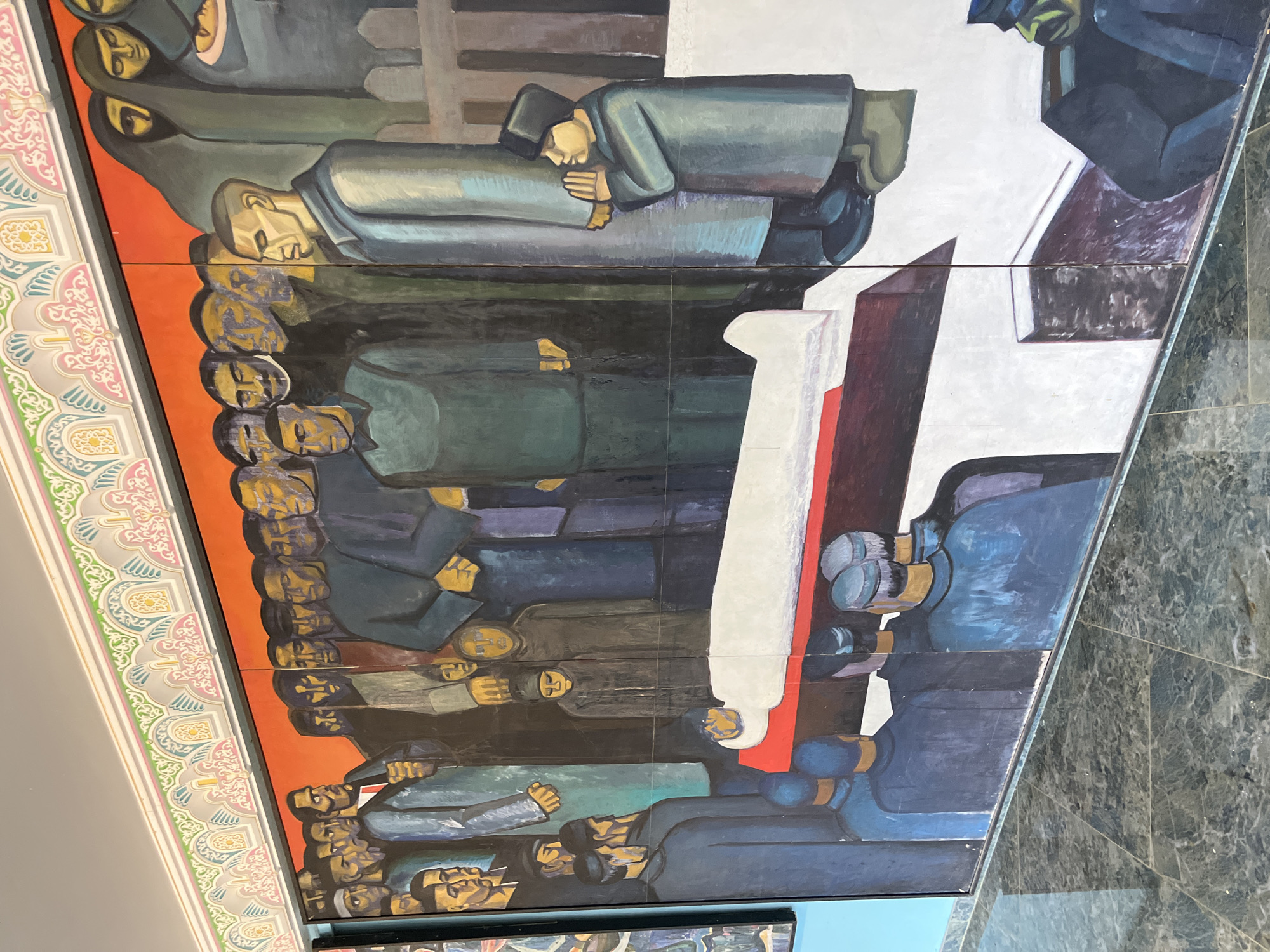 This is a photograph of a large, mural-style painting located in Kazan, Russia. The painting depicts a solemn scene with a gathering of people around a white-shrouded figure lying on a red surface, possibly a bier or coffin, in the foreground. The figures are rendered in a stylized, somewhat blocky manner with muted colors. The crowd is diverse, with individuals of varying ages and roles suggested by their clothing and postures. Some figures appear to be mourning, while others stand in solemn observation. The background of the painting features a solid orange-red sky and architectural elements that are difficult to discern clearly. Above the mural, there is decorative trim with repeating patterns in pastel colors. The floor in front of the painting is a dark, speckled marble. The mural is segmented, suggesting it is composed of multiple panels. The overall mood conveyed by the painting is one of grief, respect, and communal experience.