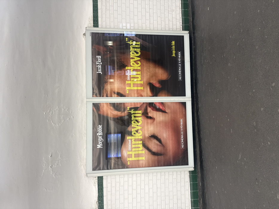 This image captures a two-panel movie poster displayed on a wall within an underground metro station in Paris, France. The poster advertises the film "Hurlevent" (Wuthering Heights), featuring close-up facial fragments. The left panel shows the lower face of a woman, identified as Margot Robbie, with closed eyes and slightly parted red lips. The right panel displays the lower face of a man, identified as Jacob Elordi, with parted lips, positioned as if about to engage in a kiss with the woman. Both panels include the text "AU CINÉMA LE 11 FÉVRIER" (In cinemas February 11th), with the right panel also stating "Jusqu'à la folie" (Until madness). The posters are mounted against a wall lined with white subway tiles and a green horizontal decorative stripe. Above the posters, the ceiling and upper wall, painted white, exhibit significant water damage and peeling paint. The floor in the immediate foreground is dark grey, textured, and appears to be a concrete platform surface. Reflections on the poster's protective covering indicate interior station lighting and what appear to be overhead electronic information displays.