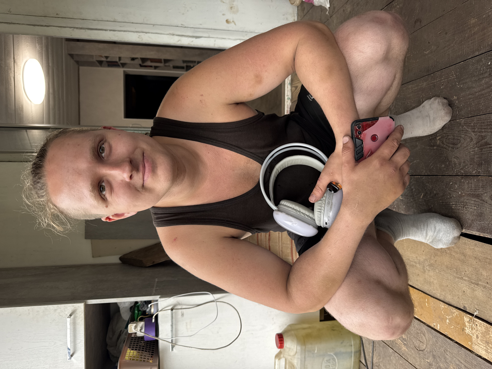 The media file shows a person crouched on a wooden floor, indoors. The person has light-colored hair pulled back, and is wearing a dark tank top and black shorts. They are holding a pink-cased smartphone in their right hand and a pair of white headphones in their left. The background includes a wall with shelves, some of which are filled with various items, and a large plastic jug with a red cap. The lighting suggests it might be daytime, possibly indoors with artificial light. The overall scene appears casual and domestic, potentially a home or workshop. The person's expression is neutral. There is no visible text in the image. The location context provided is Chaikovsky, Russia.