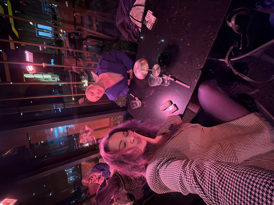 Two women sit at an outdoor table in an urban setting. One woman wears a houndstooth jacket and has reddish-purple hair. The other, across the table, is blonde and wears a blue top. The table holds glasses, an ashtray, a lighter, and what appears to be a pack of cigarettes. City lights are visible through a window in the background. The photo has a slightly blurred, reddish-purple tint.