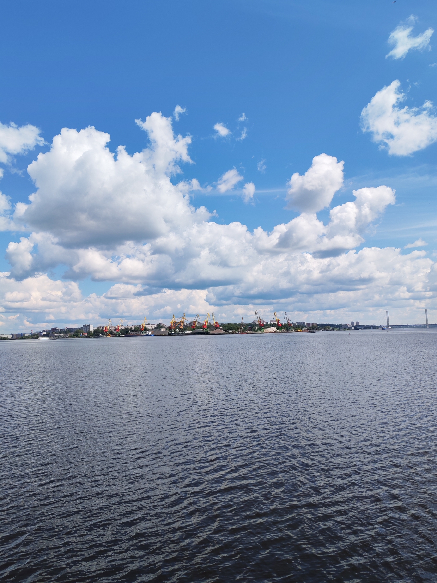 The image depicts a wide body of dark blue water with visible ripples, stretching from the foreground to a distant shoreline. On the shoreline, a cityscape is visible, characterized by buildings of various heights and a prominent industrial area with numerous tall cranes, primarily colored yellow, orange, and red. The cranes appear to be operating at a port or industrial complex. Further in the distance, a suspension bridge with two tall white pillars spans across the water, connecting to another section of the distant shore. The sky above is a vibrant blue, dotted with numerous large, fluffy white clouds, creating a dynamic and partly cloudy atmosphere. The overall scene suggests a view of a large industrial port city situated on a river or large body of water, likely Cherepovets, Russia, given the presence of the bridge and port infrastructure.