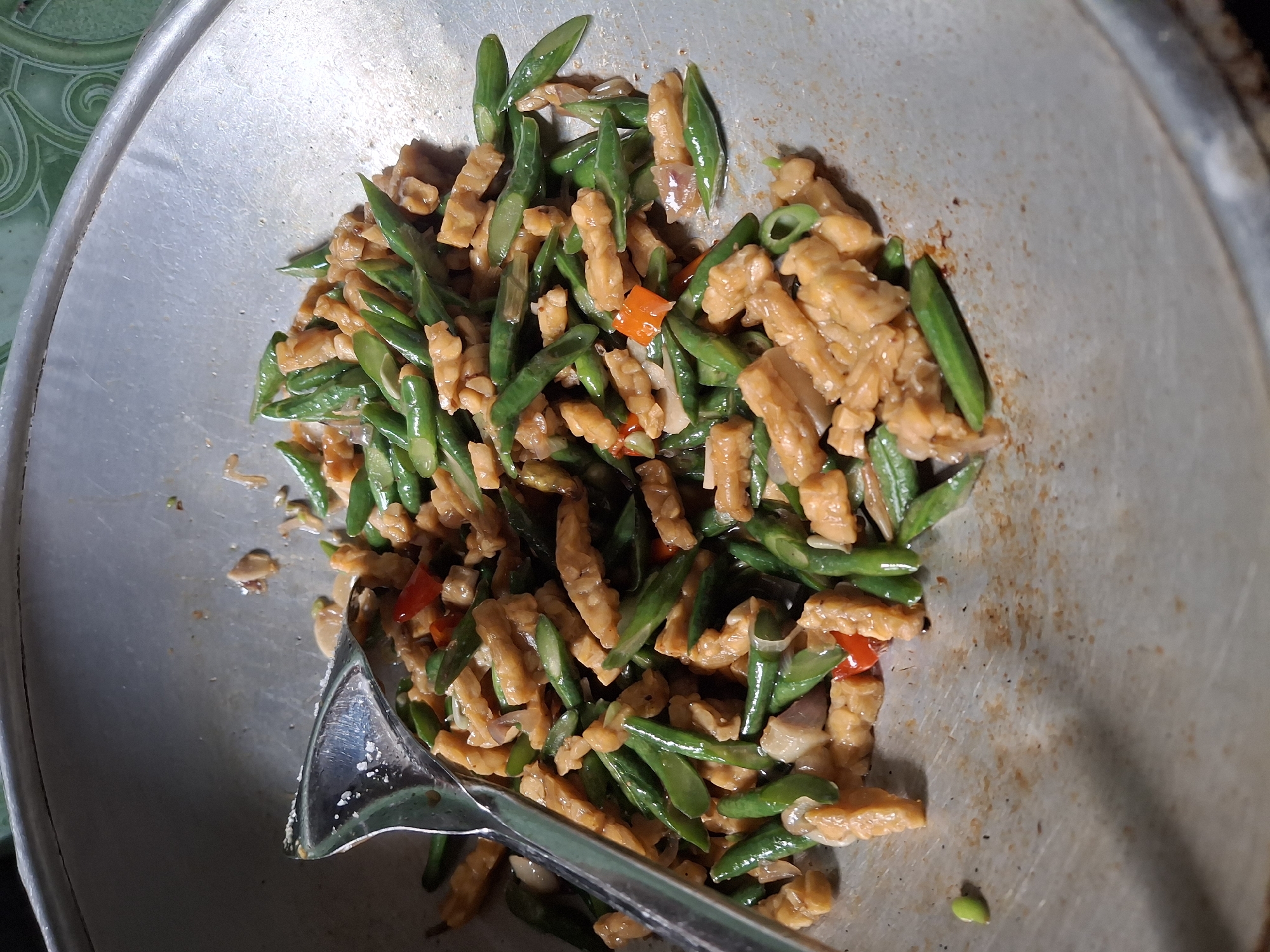 In the city of Lumajang, Indonesia, a close-up shot captures a stainless steel wok filled with stir-fried green beans and tempeh. The dish, seasoned with chilies and onions, is being mixed with a metal spatula. The wok rests on a surface, with a hint of a green tiled pattern visible in the upper right corner, suggesting an indoor or covered outdoor setting.