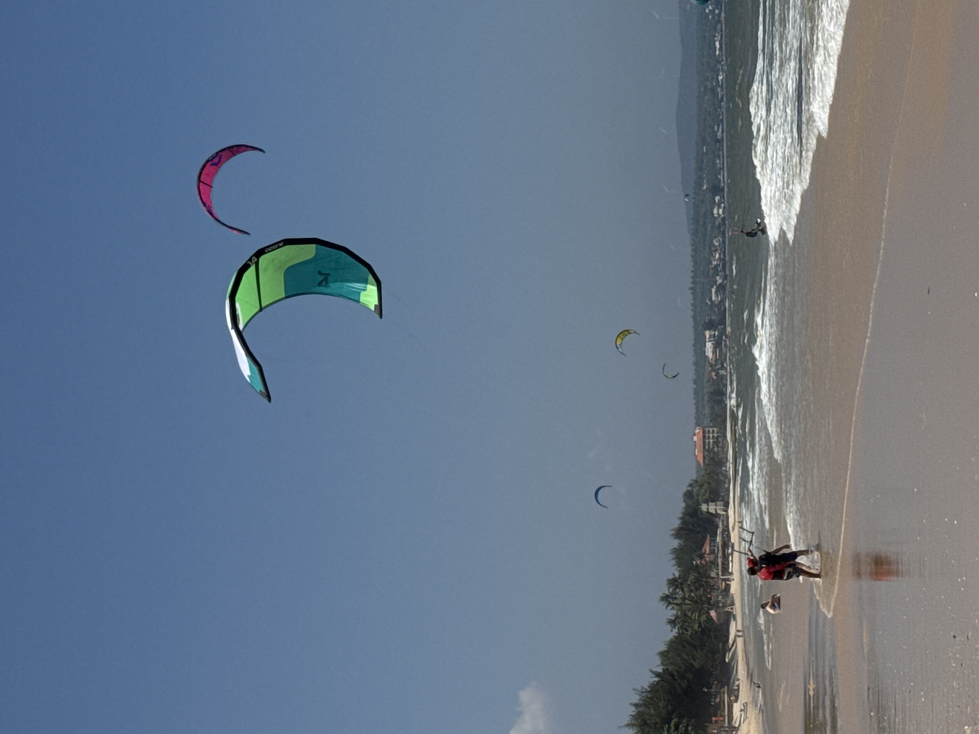 The image depicts a beach scene in Mũi Né, Vietnam, characterized by clear blue skies and a sandy coastline. The foreground shows a calm shoreline with gentle waves lapping at the sand. Two individuals are standing in the shallow water, one wearing a red and black shirt and the other a dark blue shirt, both appearing to be engaged in kiteboarding preparations or instruction. Their reflection is visible in the wet sand.
Further out in the ocean, several kitesurfers are actively engaged in their sport. A prominent green and teal kite is visible in the mid-ground, with a smaller pink kite further in the distance. Other kites, appearing as small dots against the sky, are scattered across the horizon.
The background reveals the coastal landscape of Mũi Né, with a line of trees and palm trees bordering the beach. Beyond the vegetation, buildings with red-tiled roofs are visible, suggesting a developed area. The overall impression is one of a lively beach with recreational activities taking place under a sunny sky.
