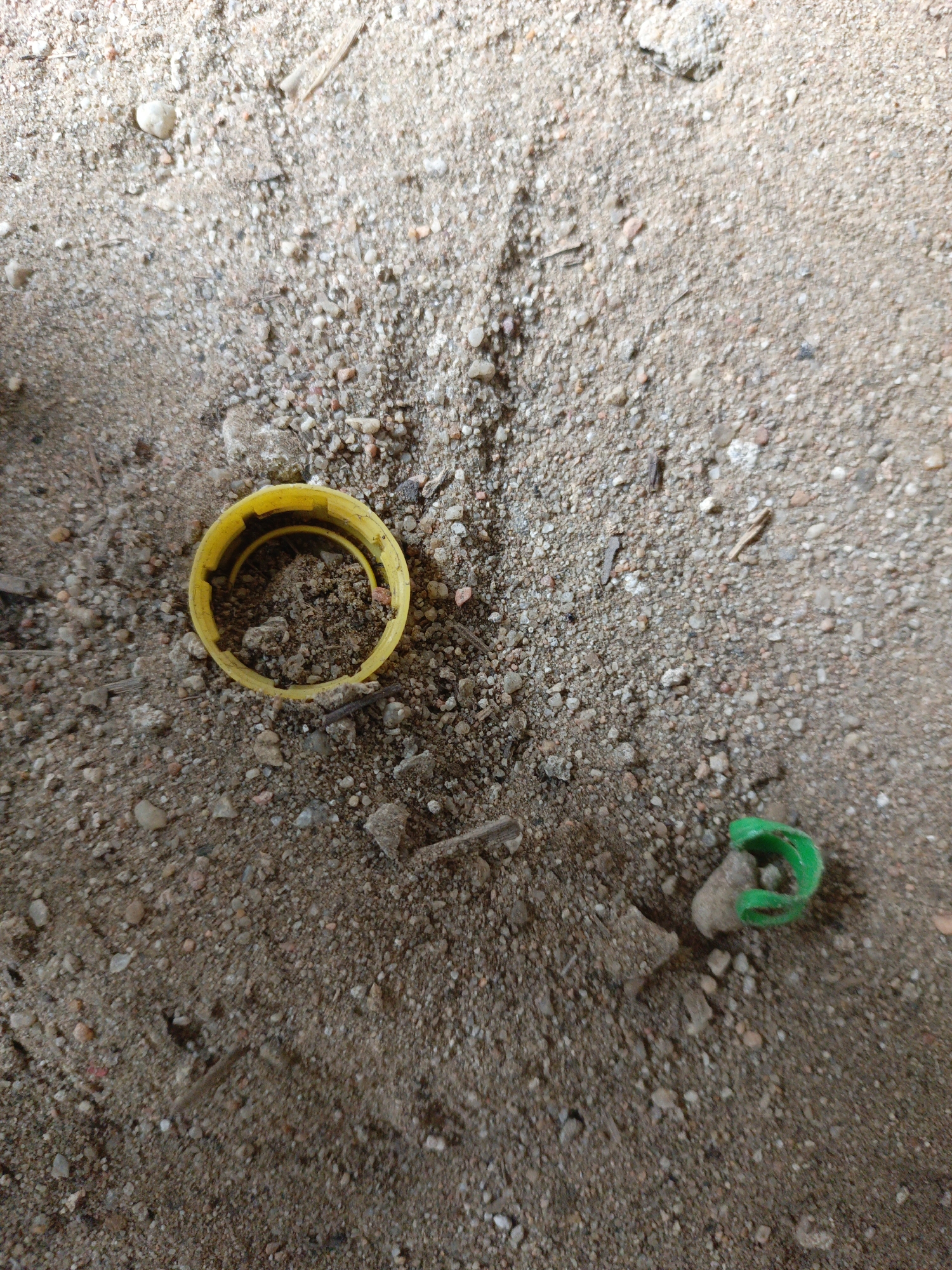 The image depicts a ground surface covered in a mix of sand, pebbles, and small debris. A yellow plastic bottle cap is lying on its side in the upper left quadrant of the frame. A green plastic clip-like object is situated in the lower right quadrant. The context or specific location within the city of Unknown, Unknown, is not discernible from the image.