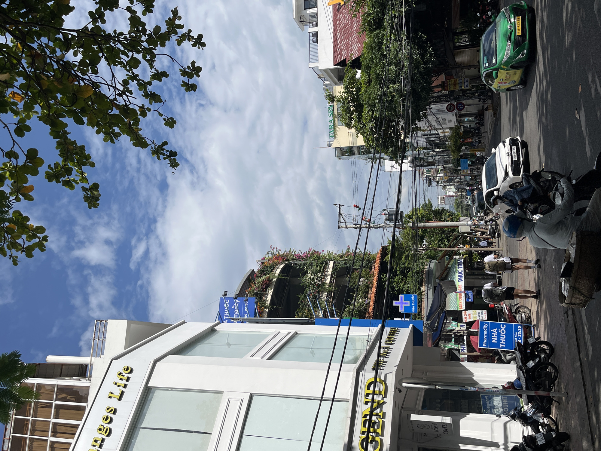 The image captures a street scene in Nha Trang, Vietnam, on a bright, cloudy day. In the foreground, a white building with large glass windows displays the signage "Legend" and "The Art Coffee." To the left of the building, several motorcycles are parked. Further down the sidewalk, a blue sign for "Pharmacity" with Vietnamese text is prominently displayed. Two individuals, each carrying a backpack, are walking away from the viewer on the sidewalk.
The street is paved with asphalt and a white car is visible in motion, followed by a green taxi with "Maxim" branding. The street is lined with buildings of varying heights, some featuring balconies adorned with lush greenery and colorful flowers. Electrical wires crisscross overhead, typical of an urban environment. Trees with green foliage are interspersed among the buildings, casting shadows on the street. In the distance, more buildings and a hint of mountainous terrain can be seen. The sky is partly cloudy with patches of blue.
