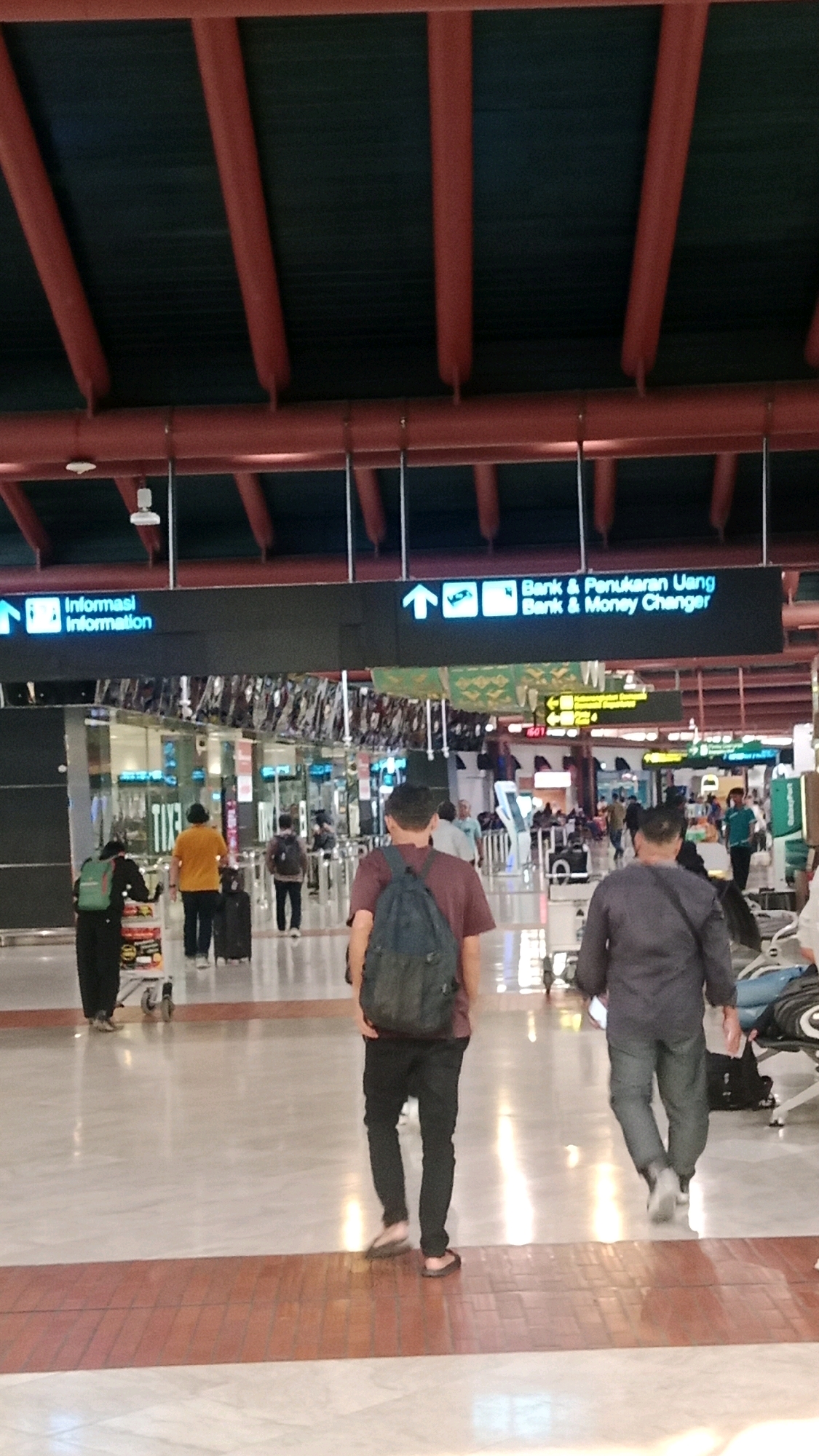 The image depicts an interior view of an airport terminal in Tangerang, Indonesia, likely Soekarno-Hatta International Airport. In the foreground, two men are walking away from the camera, one with a dark backpack and the other in a dark gray long-sleeved shirt and gray pants. The floor is polished with a light marble finish and a reddish-brown tiled strip runs horizontally across the lower part of the frame.
Above them, a large digital sign displays directions in both Indonesian and English. The left side of the sign reads "Informasi Information" with an upward arrow and an icon of an information desk. The right side shows "Bank & Penukaran Uang Bank & Money Changer" with an upward arrow and icons representing a bank and currency exchange.
Further back, other travelers are visible, some with luggage, walking towards security checkpoints or gates. The ceiling structure is prominent, with red beams and dark acoustic panels. Natural light streams in from large windows or skylights, creating reflections on the polished floor. The overall environment suggests a busy, modern airport with clear signage for passenger navigation.