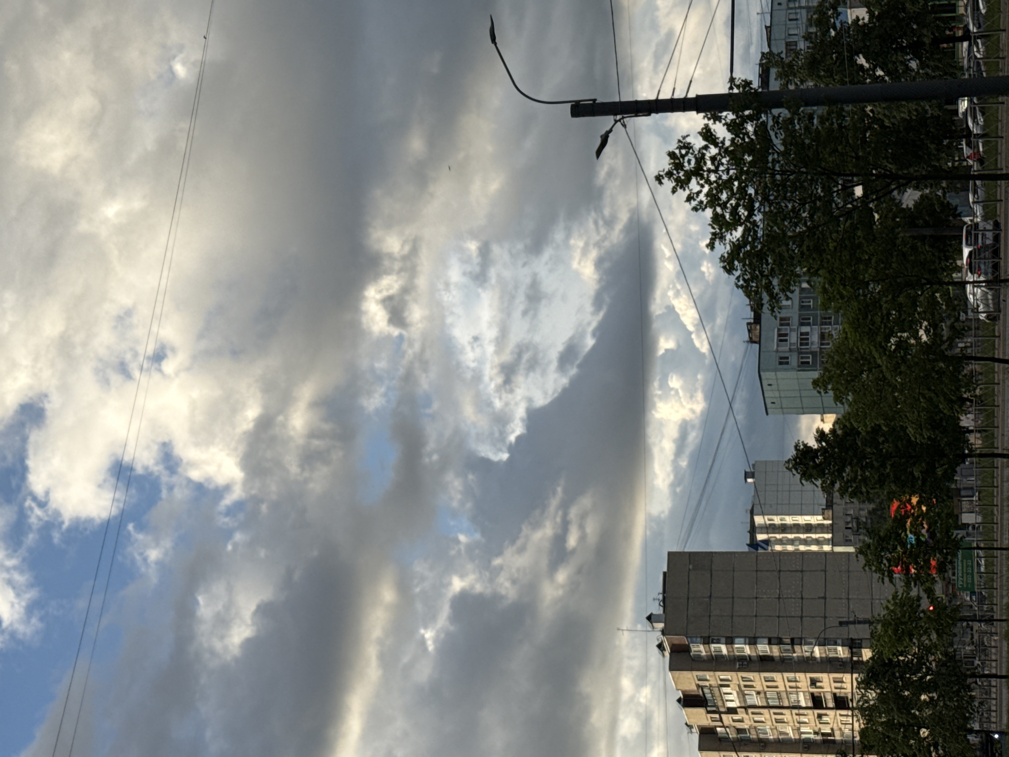 The image shows a cloudy sky over a city street in Saint Petersburg, Russia. Tall apartment buildings, trees, a lamppost, and power lines are visible. The sky is filled with dramatic clouds, with patches of blue sky peeking through. The sun is casting light on the clouds, creating a dynamic interplay of light and shadow. The scene appears to be in an urban environment, likely a residential area, given the presence of apartment buildings.