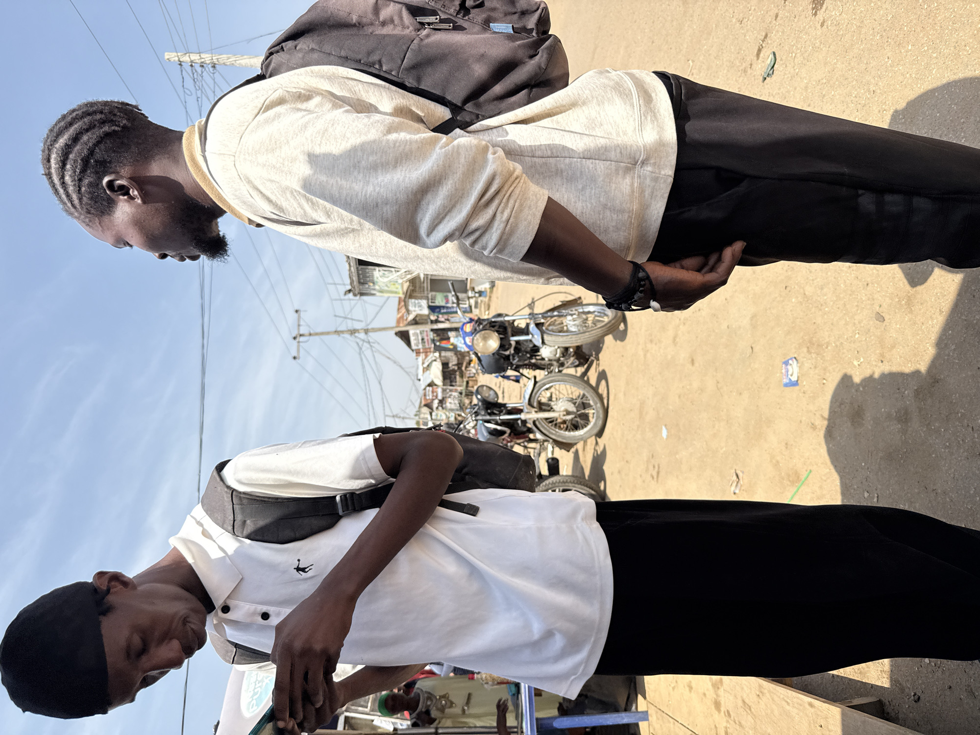 In a sun-drenched outdoor scene in Abuja, Nigeria, two young men stand facing each other in a dusty, unpaved area. The man on the left, wearing a black skullcap and a white polo shirt with a small black logo, looks down at his phone, which he holds in his hands. He has a backpack on. To his right, the second man, with braided hair and a beard, wears a light-colored long-sleeved shirt and a black backpack. He gazes off to the side. In the background, several motorcycles are parked, and some small, simple structures and utility poles are visible, suggesting a bustling, informal urban environment, possibly a market or roadside area. The ground is rough, with some scattered debris.