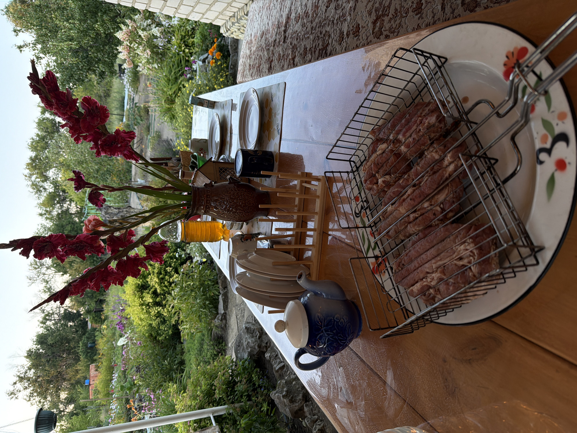 A low-angle shot captures a wooden table laden with a meal outdoors. In the foreground, a grill basket filled with raw sausages rests on a decorative plate. Behind the sausages, a blue teapot with a white lid sits on the table, next to a wooden dish rack holding several plates. Further back, a vase of vibrant red gladioli stands tall, accompanied by a bottle of amber liquid and other tableware. The table is set against a backdrop of a lush, blooming garden with a brick wall visible to the right. The scene suggests a pleasant outdoor dining experience, possibly in a residential area. There are no people visible in the image. The image likely depicts a summer scene in Kamyshin, Russia, given the presence of a well-maintained garden and outdoor dining setup typical of dacha culture.