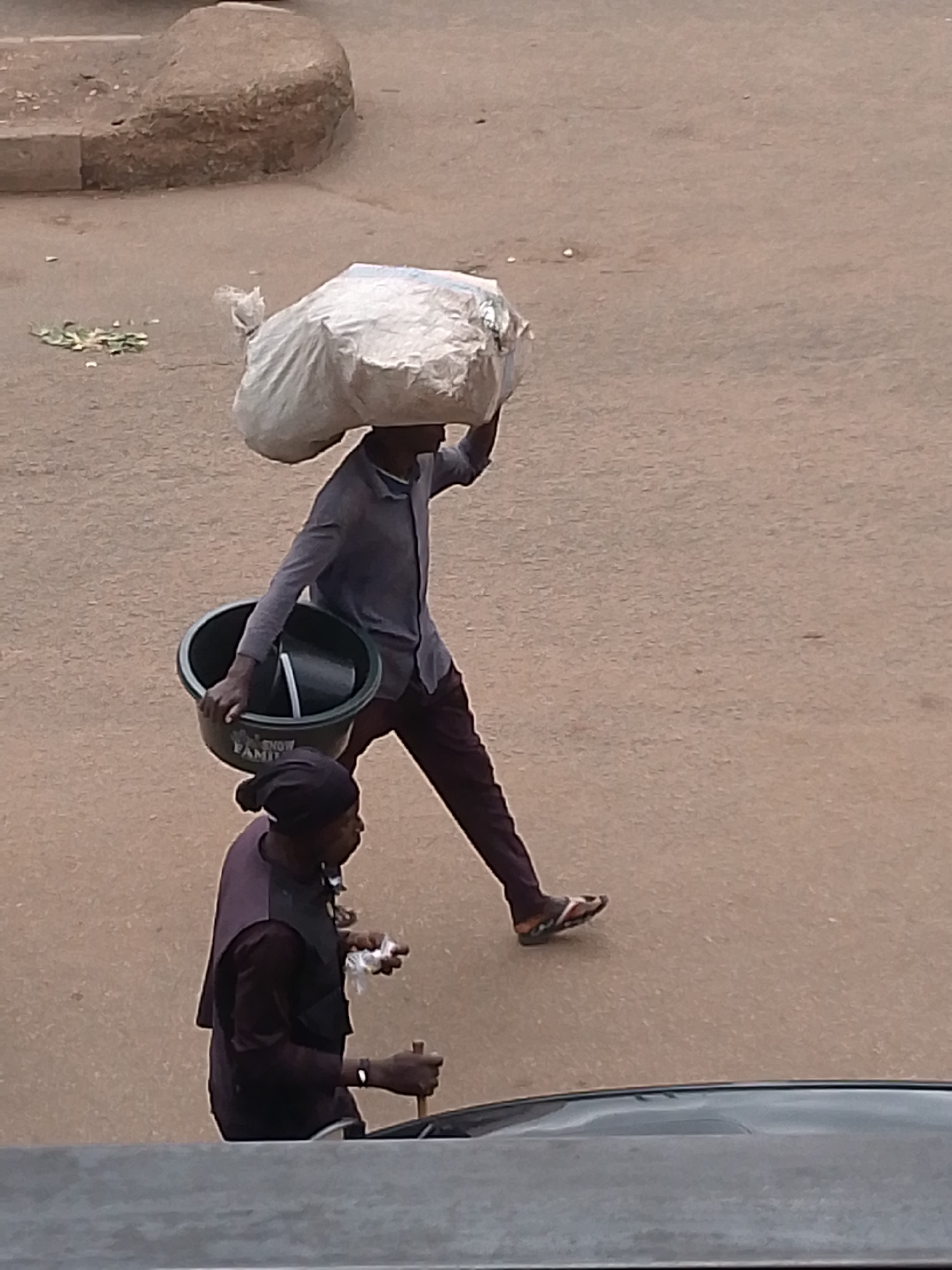In Bukuru, Nigeria, a person walks along a paved surface, carrying a large, white plastic bag balanced on their head. The person is wearing a blue collared shirt, dark pants, and sandals. They are also holding a large, dark basin with their left hand. Below and slightly behind the first person, another individual is visible, wearing a dark head covering and a dark vest over a long-sleeved shirt. This second person appears to be holding a small white object in their right hand and a wooden stick in their left hand, near a dark object that could be a vehicle. The environment appears to be an outdoor, urban setting, with a sandy or dusty ground.