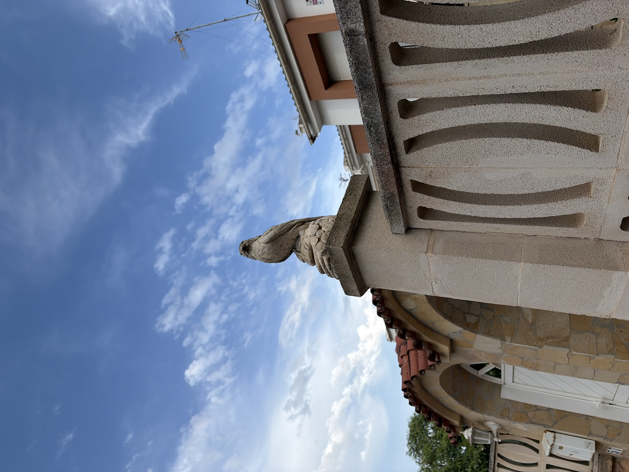 A low-angle shot captures a stone statue of a bird perched on a pedestal atop a stone pillar, against a backdrop of a blue sky with scattered white clouds. In the middle ground, a section of a fence with curved openings runs horizontally across the frame, and behind it are houses with terracotta tiled roofs and white and orange facades. To the left, an arched entryway with stone cladding and a white door is visible. The scene is outdoors, with hints of greenery from trees on the far left.