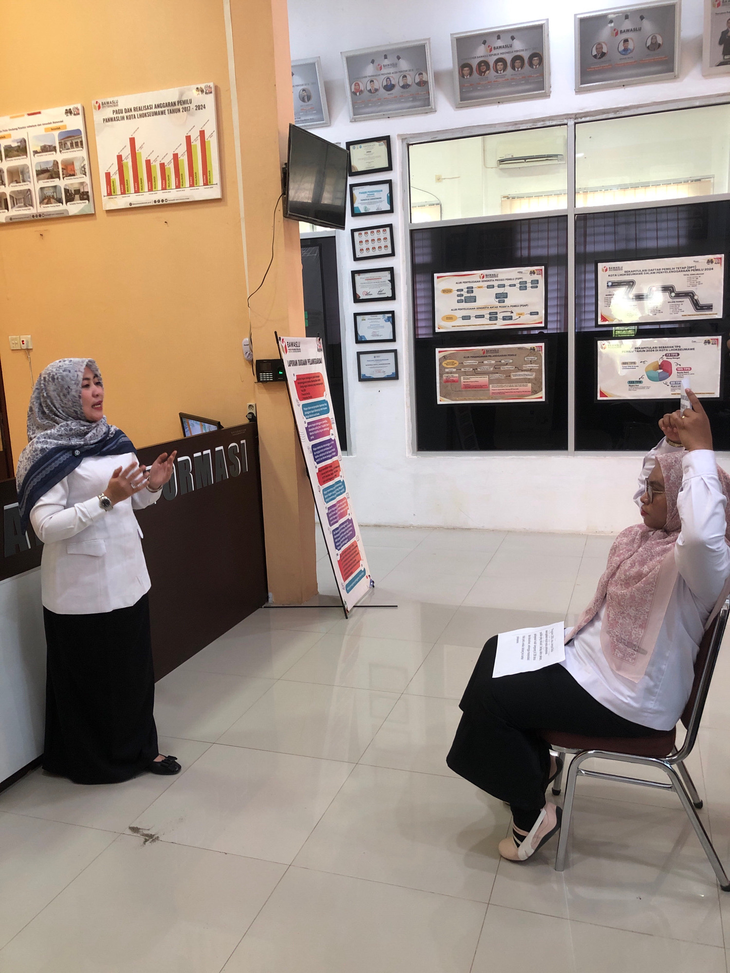The image depicts an indoor scene, likely an office or meeting room, situated in Lhokseumawe, Indonesia. In the foreground, two women are present. One woman, wearing a white long-sleeved blouse and a black skirt, stands and gestures with her hands as if speaking. She has a hijab with a grey and blue pattern. Behind her is a counter with "INFORMASI" printed on it. The second woman is seated in a chair, wearing a light pink hijab, glasses, and a white top with a pink patterned overlay. She is holding a document and pointing upwards with her right hand. The environment features tiled flooring and walls adorned with framed posters and charts. The posters on the wall appear to be related to elections and government administration, indicated by the "BAWASLU" (Badan Pengawas Pemilihan Umum - Election Supervisory Body) logo present on several of them. The presence of election-related materials strongly suggests the location is an office of the Election Supervisory Body in Lhokseumawe.