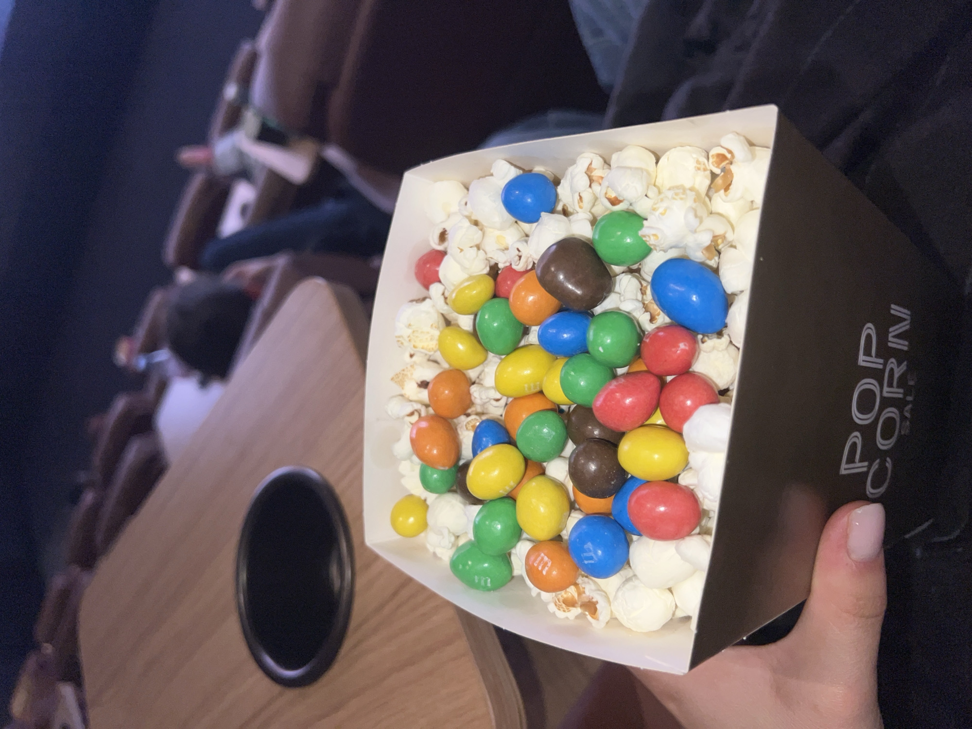 The image shows a box of popcorn mixed with M&Ms, held by a person with light pink nail polish. The box is predominantly dark with white "POPCORN" lettering visible. The popcorn is white and fluffy, and the M&Ms are in various colors including blue, green, yellow, orange, red, and brown.
The scene appears to be inside a movie theatre, as rows of seats are visible in the background. A wooden table with a cup holder is also present.
There is no information in the image to indicate the location is Paris, France.