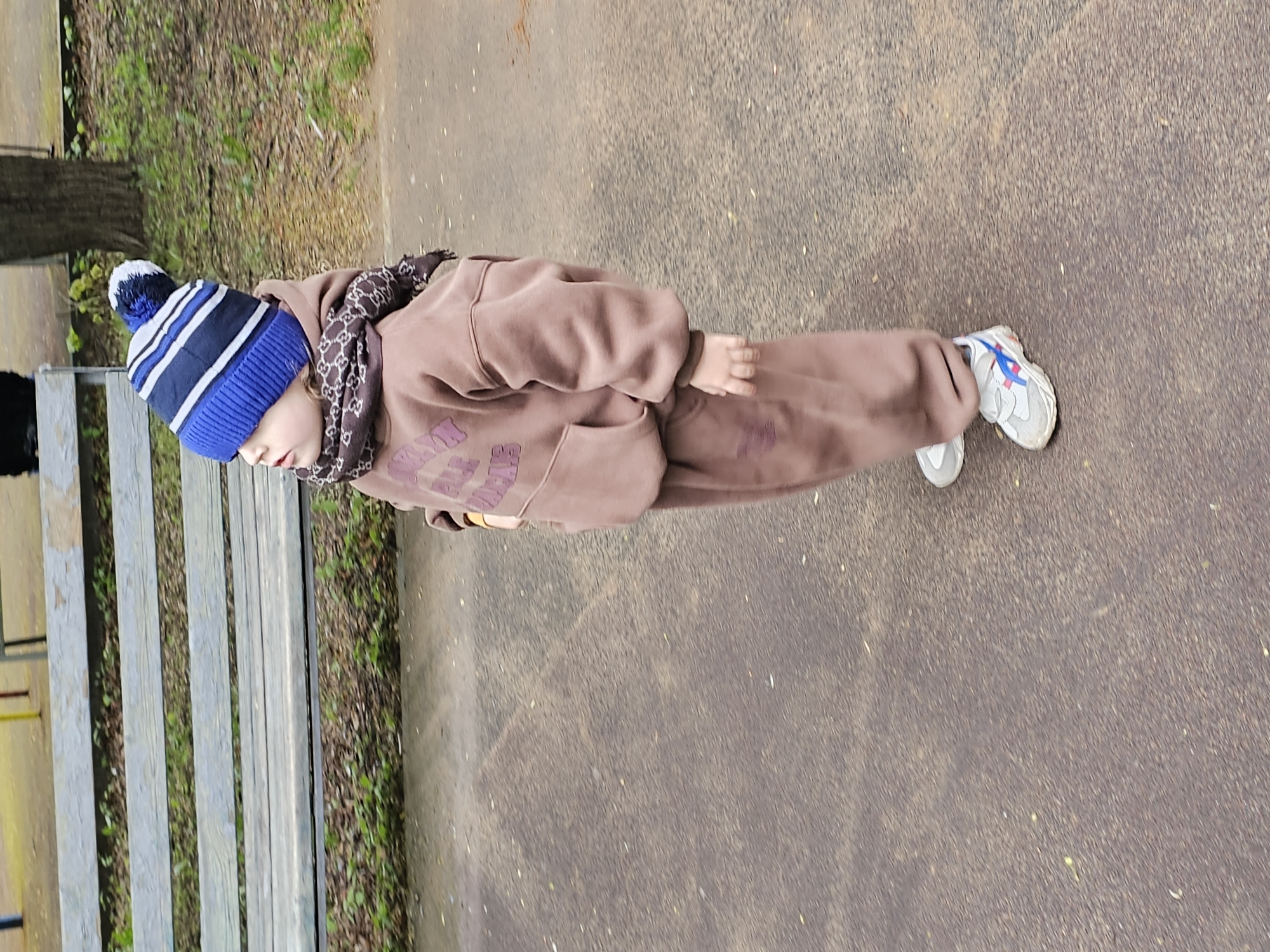 A young person stands on a concrete surface in what appears to be a park or outdoor area. The person is dressed in a brown tracksuit with a patterned scarf and a blue and white striped beanie. They are wearing white and blue sneakers. A wooden bench is visible to the left of the person. The concrete surface is speckled with debris and natural materials. The background includes trees and foliage. The location is Khimki, Russia.
