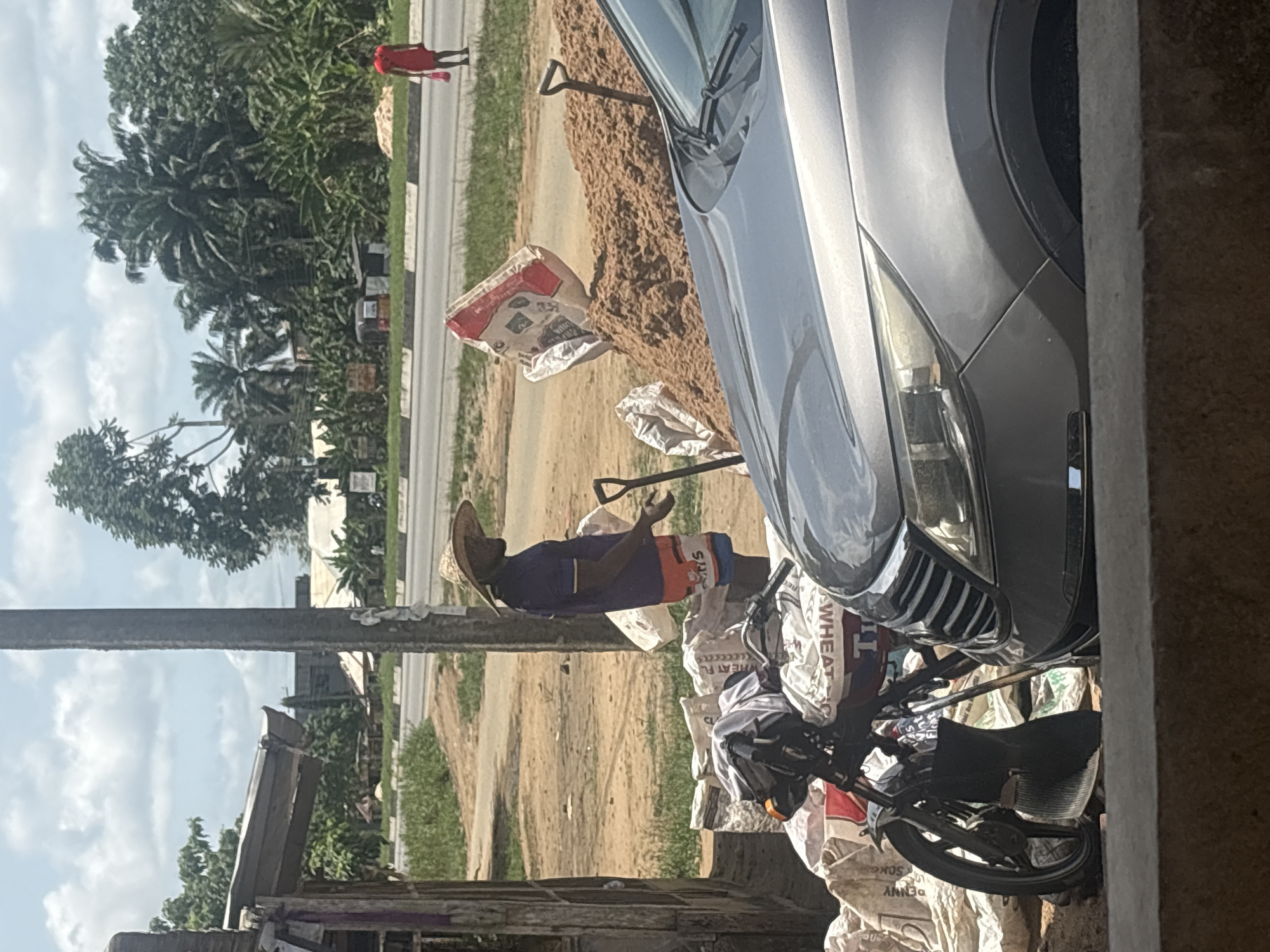 The image shows an outdoor scene in Uyo, Nigeria. A dark-skinned man wearing a straw hat and blue shirt, and orange shorts is standing next to a pile of sand and several bags. The bags appear to contain grain, some are labeled "WHEAT". A shovel leans against the sand pile. A gray car is parked nearby. A black motorbike is parked in front of the bags, and near the front of the car. In the background, a road is visible, with a person wearing a red shirt walking along it. There are trees and buildings in the background, and a power pole and old wooden structure in the foreground. The sky is partly cloudy.