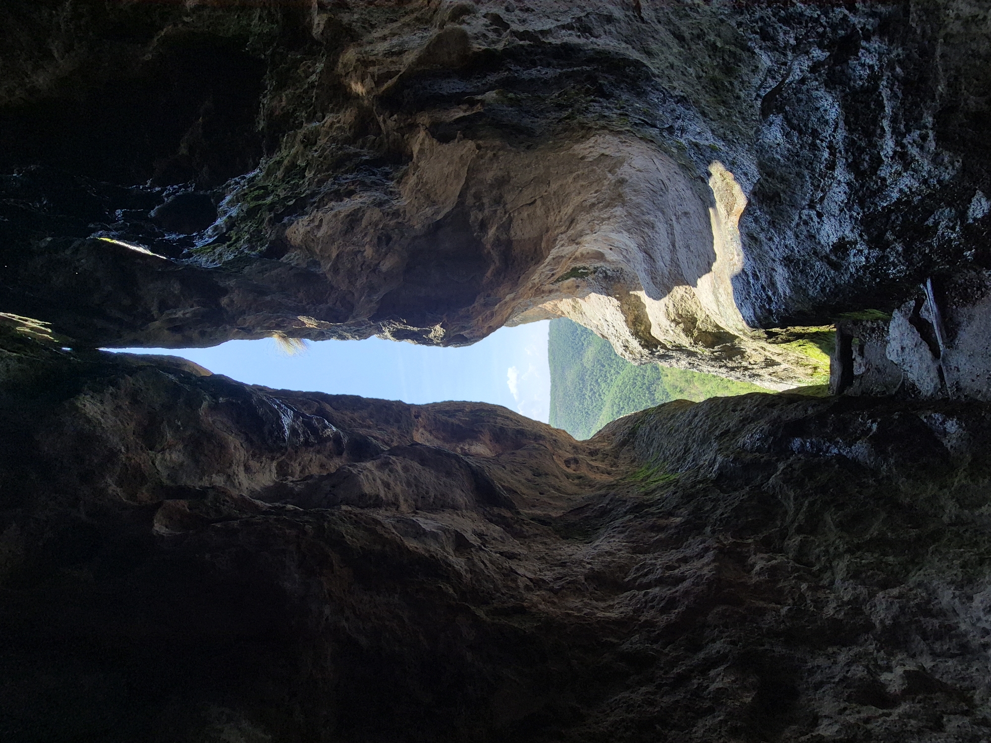 The image captures a perspective from within a deep, natural rock fissure, located near Glavatartsi, Bulgaria, looking outward through a narrow vertical opening. The internal rock walls, on both the left and right, are massive, irregular, and heavily textured, displaying a range of dark brown, grey, and shadowed tones indicative of natural erosion and weathering. Patches of green moss cling to the damp surfaces, particularly visible on the lower right wall.
Daylight streams primarily through the central vertical aperture, illuminating the outer edges of the rock walls and contrasting sharply with the dimly lit, darker recesses of the interior. On the right-hand wall, a section of rock is directly exposed to sunlight, appearing brighter and more beige-toned, creating a prominent shadow directly below it. The ground at the base of the fissure is a narrow, uneven path composed of small rocks, dirt, and some broken woody debris, leading towards the exterior.
Through the tall, narrow opening, a bright, clear blue sky occupies the upper portion of the distant view, with a small, wispy white cloud visible slightly to the right of center. Below the sky, a panoramic view of distant rolling hills and mountains covered in dense green foliage is visible under full daylight. The scene is devoid of human figures, animals, or man-made structures.