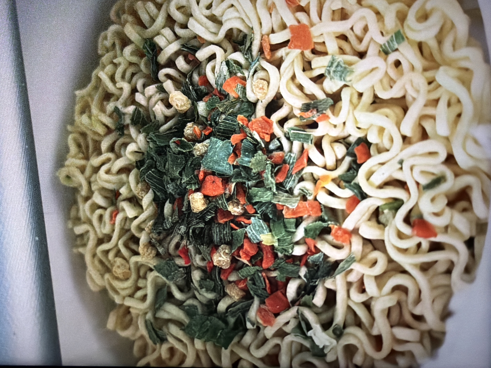 A close-up, overhead shot shows a package of instant ramen noodles. The noodles are coiled and dry, with a topping of dehydrated vegetables, including green pieces that appear to be chives or scallions, and small orange pieces that are likely carrots. There are also some lighter colored, irregularly shaped pieces that may be dried mushrooms or other seasoning. The noodles fill most of the frame, spilling out of a white cardboard container, the edge of which is visible at the top and bottom of the image. The lighting is bright and somewhat diffused, highlighting the textures of the noodles and dried vegetables. The location in Krasnodar, Russia is not discernible from the image.