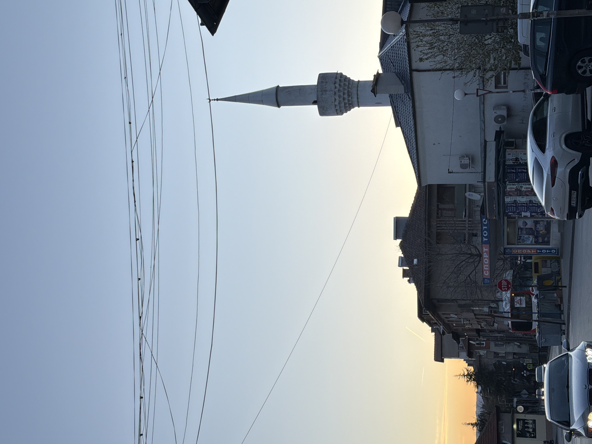 The image shows a street scene in Blagoevgrad, Bulgaria. A minaret belonging to the Orta Dzhamiya Mosque is visible on the right side. Power lines are seen on the left side of the image. The buildings on the right side of the image appear to be a mix of residential and commercial, with signs in Bulgarian visible. Cars are parked along the side of the street and driving by, including a white SUV and a dark colored sedan. A stop sign is also visible. The sky is a light blue with a hint of orange near the horizon, suggesting either sunrise or sunset.