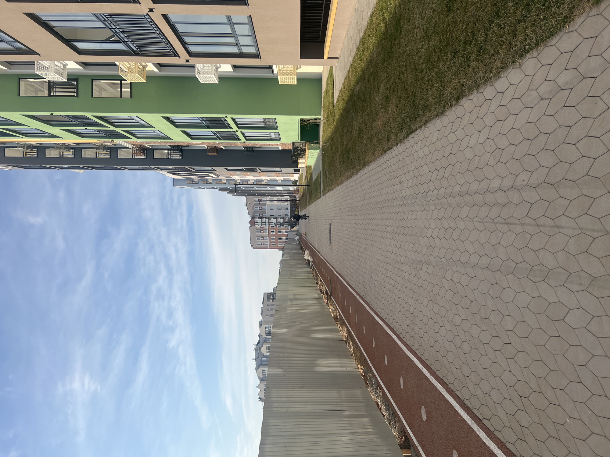 The image depicts a street scene in Pervomaysky, Russia. Buildings of varying heights and colors line one side of the street. A paved walkway with a running track runs along the buildings. Beyond the running track, there is a low metal fence/wall. The sky is blue with scattered clouds. Two individuals are visible in the distance on the walkway. Vegetation (grass) is visible between the buildings and the paved walkway. The perspective is at ground level, looking down the street.
