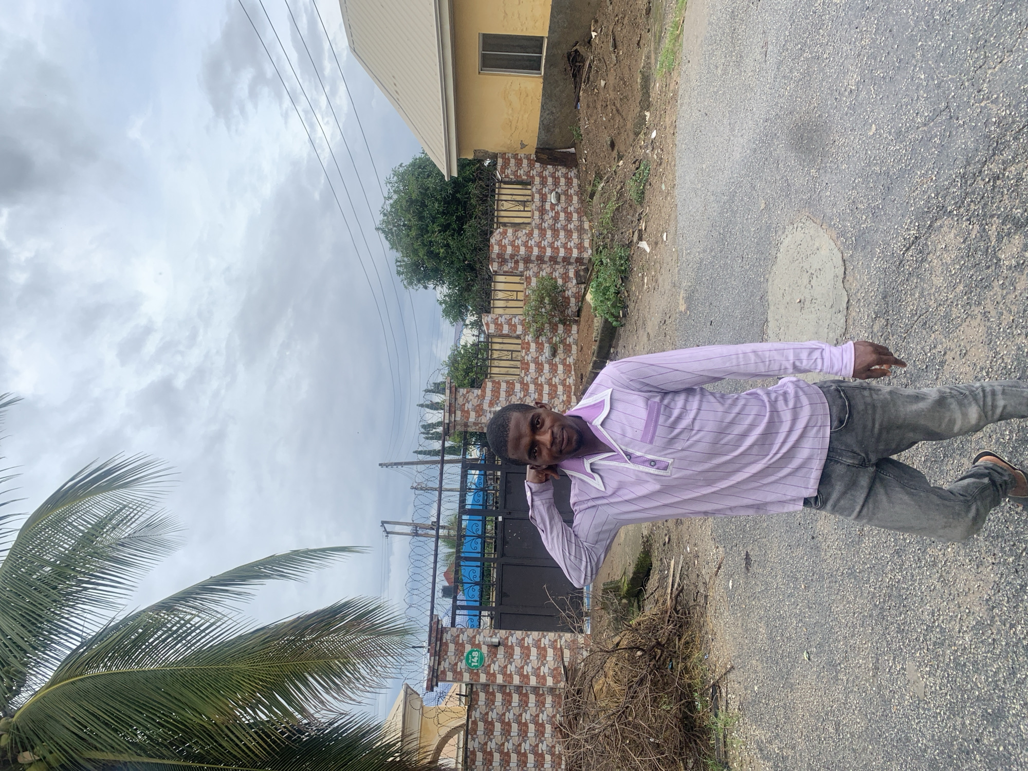 A medium shot captures a Black male, appearing to be in his late 20s or early 30s, standing outdoors in an urban environment, likely Abuja, Nigeria. He is facing the camera with his left hand touching the back of his head. He has short black hair and a short beard. He is wearing a light purple collared shirt with thin vertical pinstripes and a single chest pocket, and dark gray, faded jeans. He has brown sandals on his feet.
The man is positioned on a gravelly asphalt road. Behind him is a decorative brick wall topped with barbed wire fencing. A large black metal gate with ornate scrollwork is visible within the wall, and beyond the gate, a bright blue structure, possibly a swimming pool, is partially visible. To the left of the wall, a large pile of dried branches and debris sits on the ground. To the right of the man and wall, a yellow building with a cream-colored roof and a single window is visible. The sky above is overcast with gray clouds. The overall scene suggests a residential or semi-commercial area.