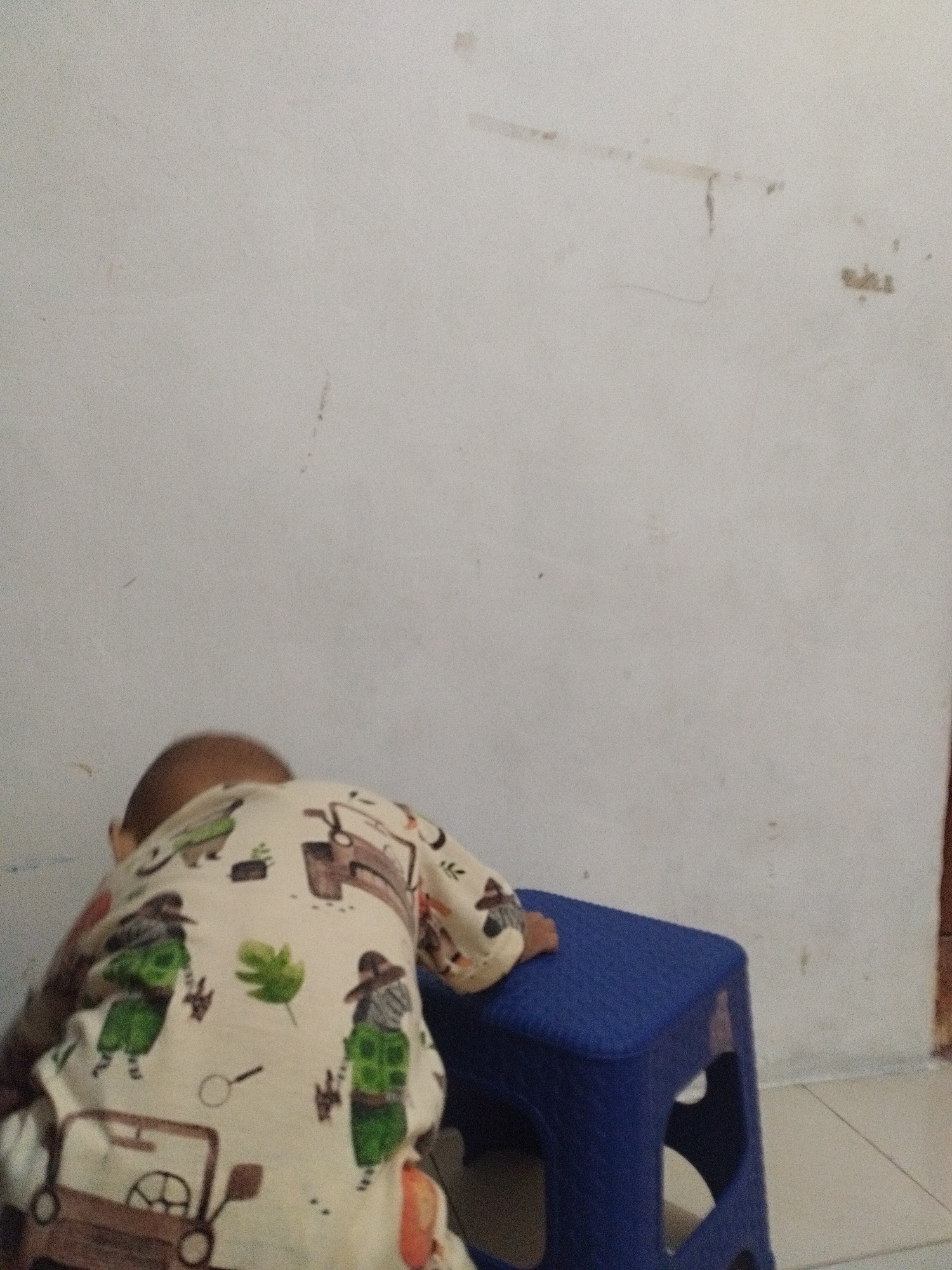 In Surabaya, Indonesia, a young child wearing pajamas with a print of cartoon characters and a toy jeep is bent over a blue plastic stool. The child's back is to the camera, and their head is down. The background is a white wall with some faint marks and shadows. The floor is tiled with light-colored tiles. The scene appears to be indoors.