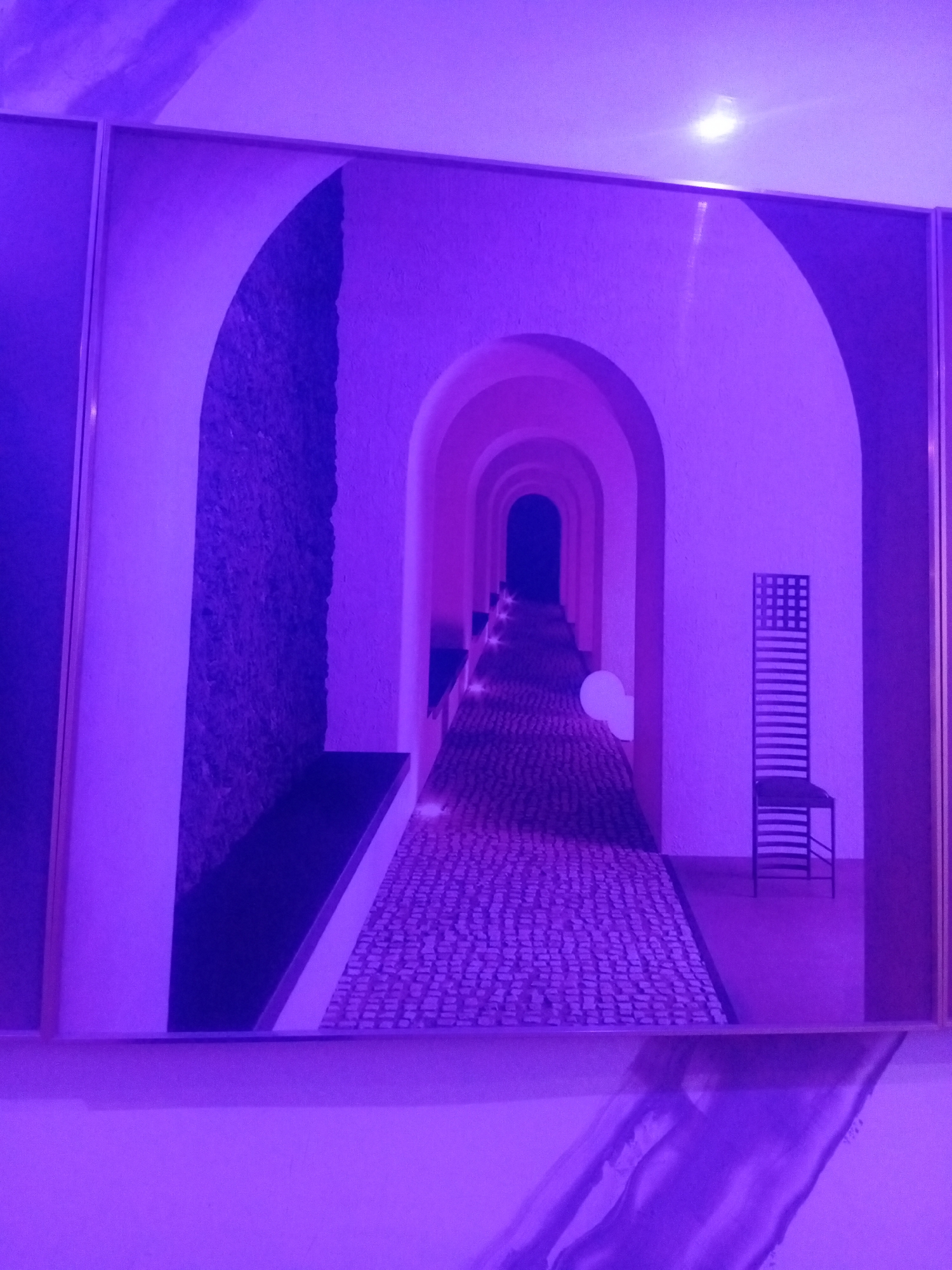 The image shows a framed artwork depicting a long, arched hallway with a cobblestone floor. The artwork is illuminated by purple lighting, which casts a strong violet hue over the entire scene. In the foreground, to the right, a tall, narrow chair with a grid-like back and horizontal slats is visible. The hallway recedes into the distance, with a series of arches creating a sense of depth and perspective. The walls of the hallway appear to be textured, with one section to the left covered in a dark, rough material that resembles moss or a rough-textured wall covering. A white, spherical object is subtly placed in the middle of the hallway, further back. The overall impression is one of an architectural study, likely an artist's interpretation of an interior space. The specific location in Sabon Gari East, Nigeria, is not discernible from the visual information present in the artwork itself.