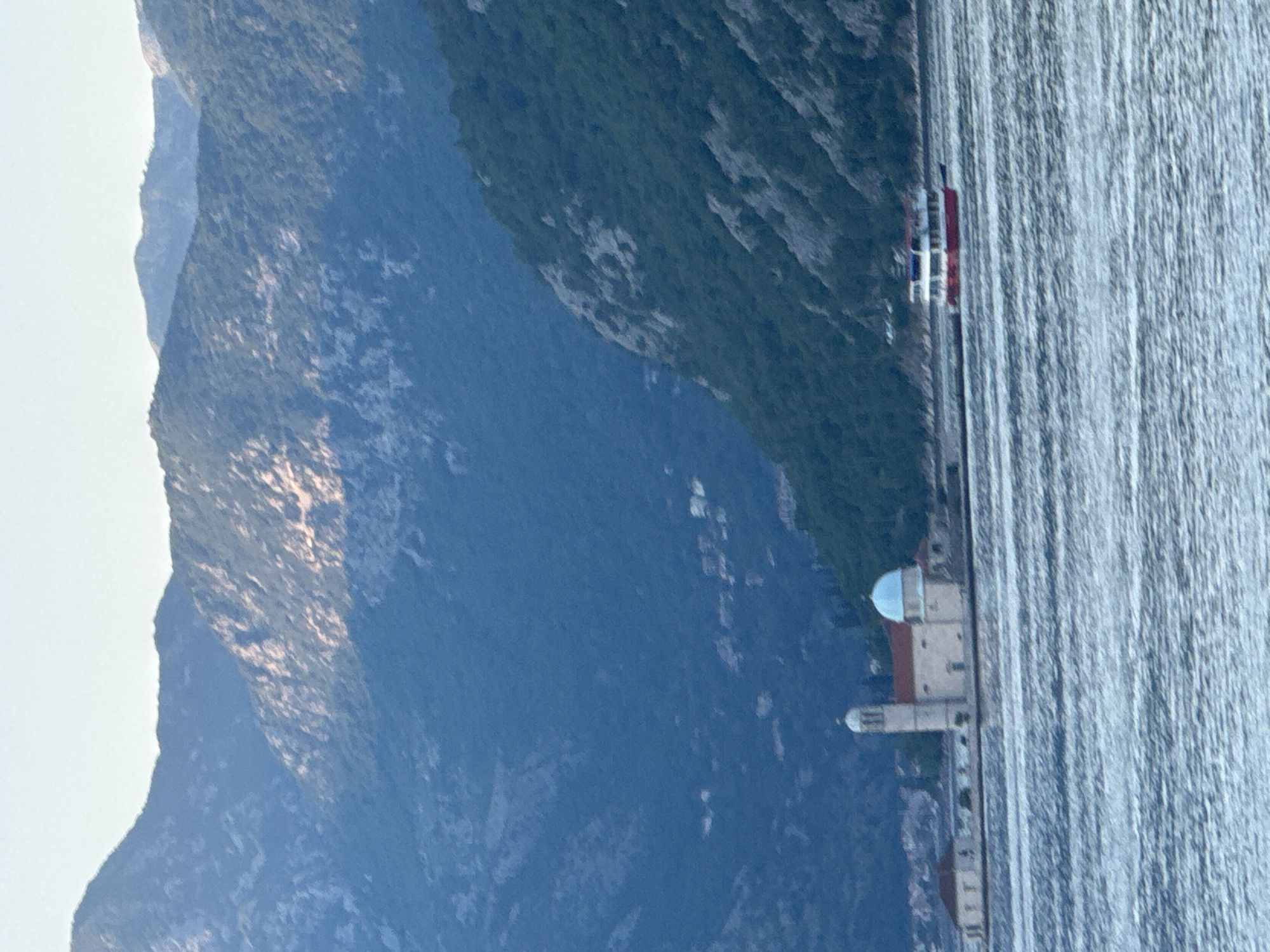 A panoramic view shows the mountains of Đurići, Montenegro, overlooking the waterfront and the shimmering sea. A church with a distinctive dome sits near the shore. A boat is navigating the waterway.