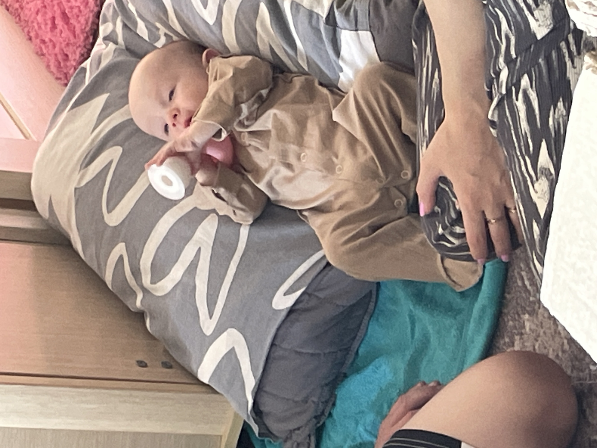 A baby in a tan jumpsuit lies on a grey pillow with a white wavy pattern, holding a baby bottle. An adult's hand with pink nail polish and a gold ring rests near the baby, adjacent to a patterned garment. The scene includes a blue blanket and part of a bed. The location is identified as Perm, Russia.