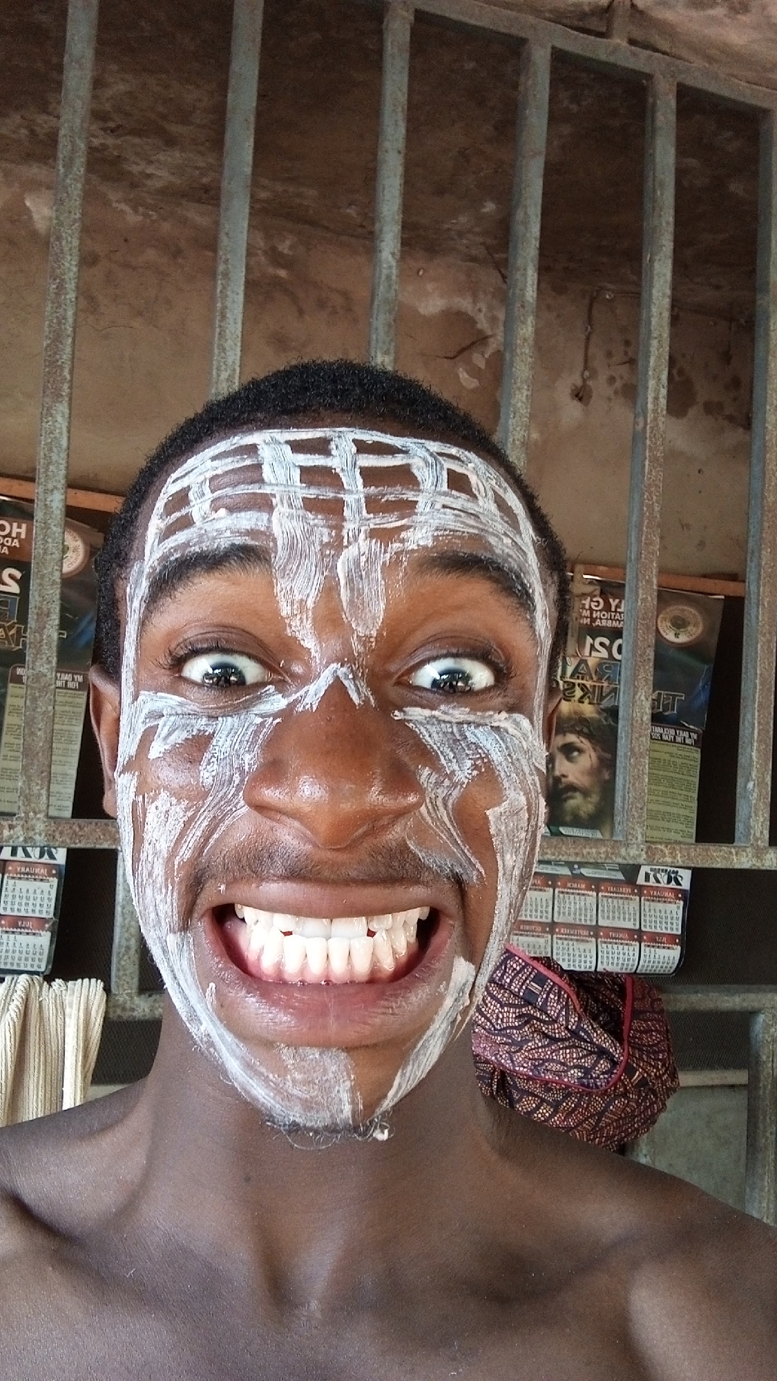 A close-up portrait features a person, likely a young man, with dark skin and short dark hair, looking directly at the camera. His face is extensively painted with white patterns, including grid-like lines on his forehead, streaks across his cheeks and around his eyes, and designs on his nose and chin. He displays a wide, open-mouthed grin, revealing bright white teeth, and his eyes are wide open and intense.
Behind the person, several vertical metal bars are visible, suggesting a window or gate. Through these bars, posters and calendars are partially seen. On the left, fragments of text include "HC", "GA", "S", "PLA OVICA" (reversed), "YM" (reversed), "M HOITAS" (reversed), and "НИ АЯЗМА" (reversed). On the right side of the person's head, a poster features a depiction of Jesus Christ. Below this, parts of calendars are visible, displaying text like "YRAUKAL" (reversed) and "YRAUWAL" (reversed). A patterned, reddish-brown fabric object, possibly a small bag or cloth, hangs behind his right shoulder. The background wall behind the bars appears to be a rough, light brown surface with some marks.
The person's expression conveys an exaggerated sense of excitement, joy, or playfulness.