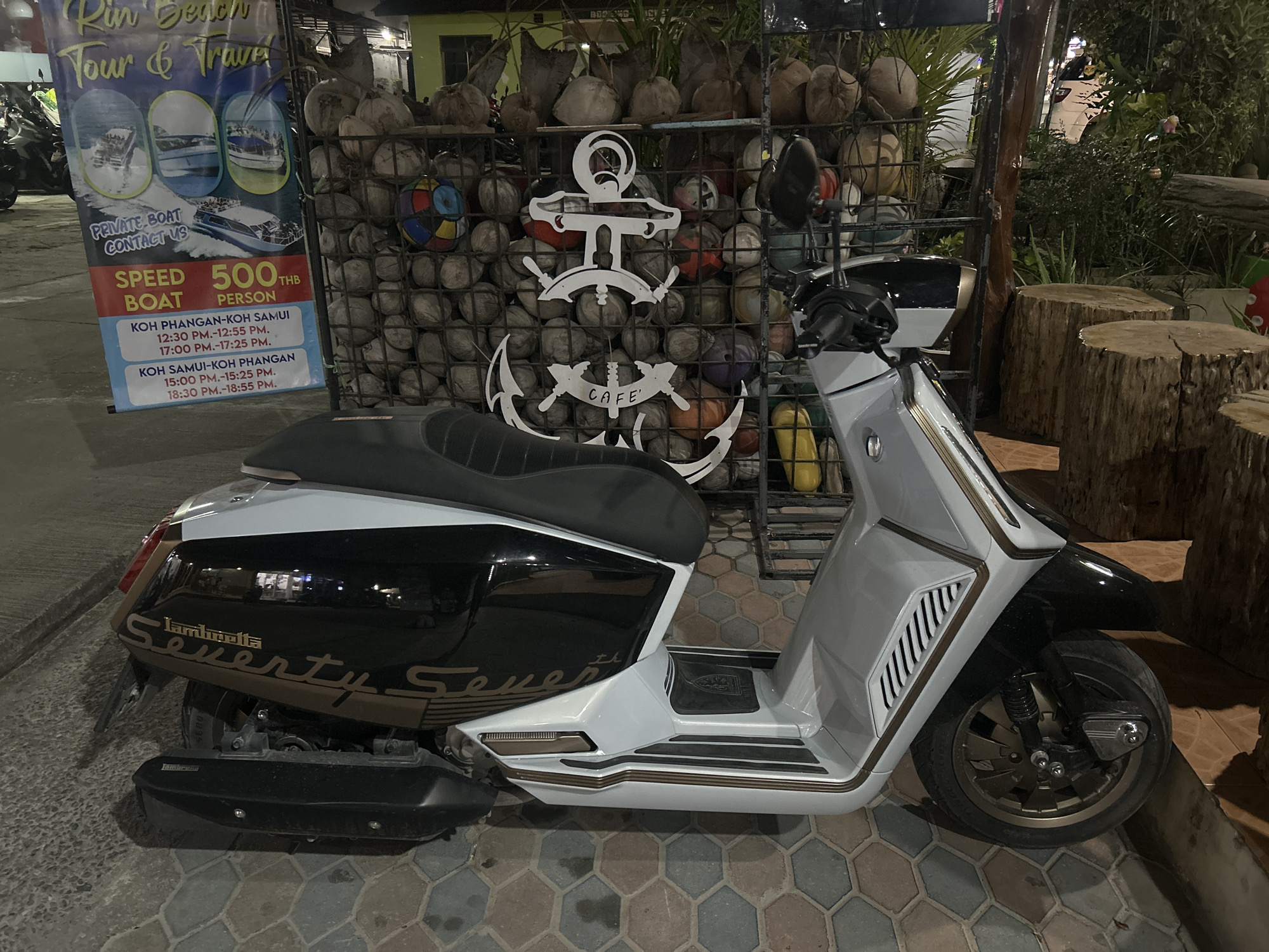 The image shows a Lambretta Seventy Se7en scooter parked in what appears to be an outdoor area in Ban Haad Rin, Thailand. The scooter is primarily white and black with gold trim. Behind the scooter is a metal grid filled with coconuts and various spherical objects. An anchor with the word "CAFE" on the bottom and a Rin Beach Tour & Travel sign are affixed to the grid. Several tree stump-like structures are located to the right of the scooter. The ground is paved with hexagonally shaped tiles. The scene seems to be lit artificially and has a mix of natural elements and urban features.