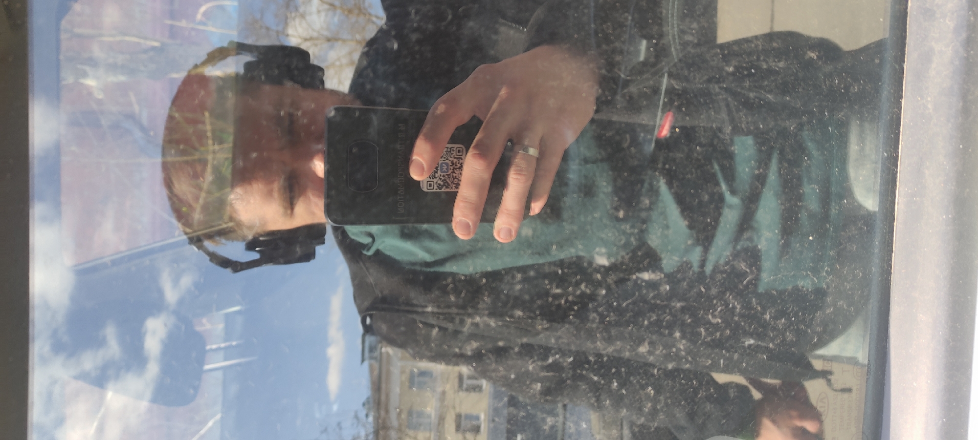The image shows a person taking a selfie in a reflective window, likely a car window. The reflection shows the person's face, wearing headgear of some kind, and the hand holding the phone. The phone's screen displays a QR code. On the hand holding the phone, a ring is visible. The background reflection shows a building, trees, and the sky. A reflection of the person's jacket can be seen. At the bottom of the frame is a sticker that reads "KIA" with text in Russian. Based on the text visible on the sticker ("2071 REАБ 50", "2071 REAB", "КІА"), this is the inspection sticker of the vehicle. The city is likely Fryazino, Russia, as the Russian text confirms it is in Russia. The window is dirty with dust and debris.