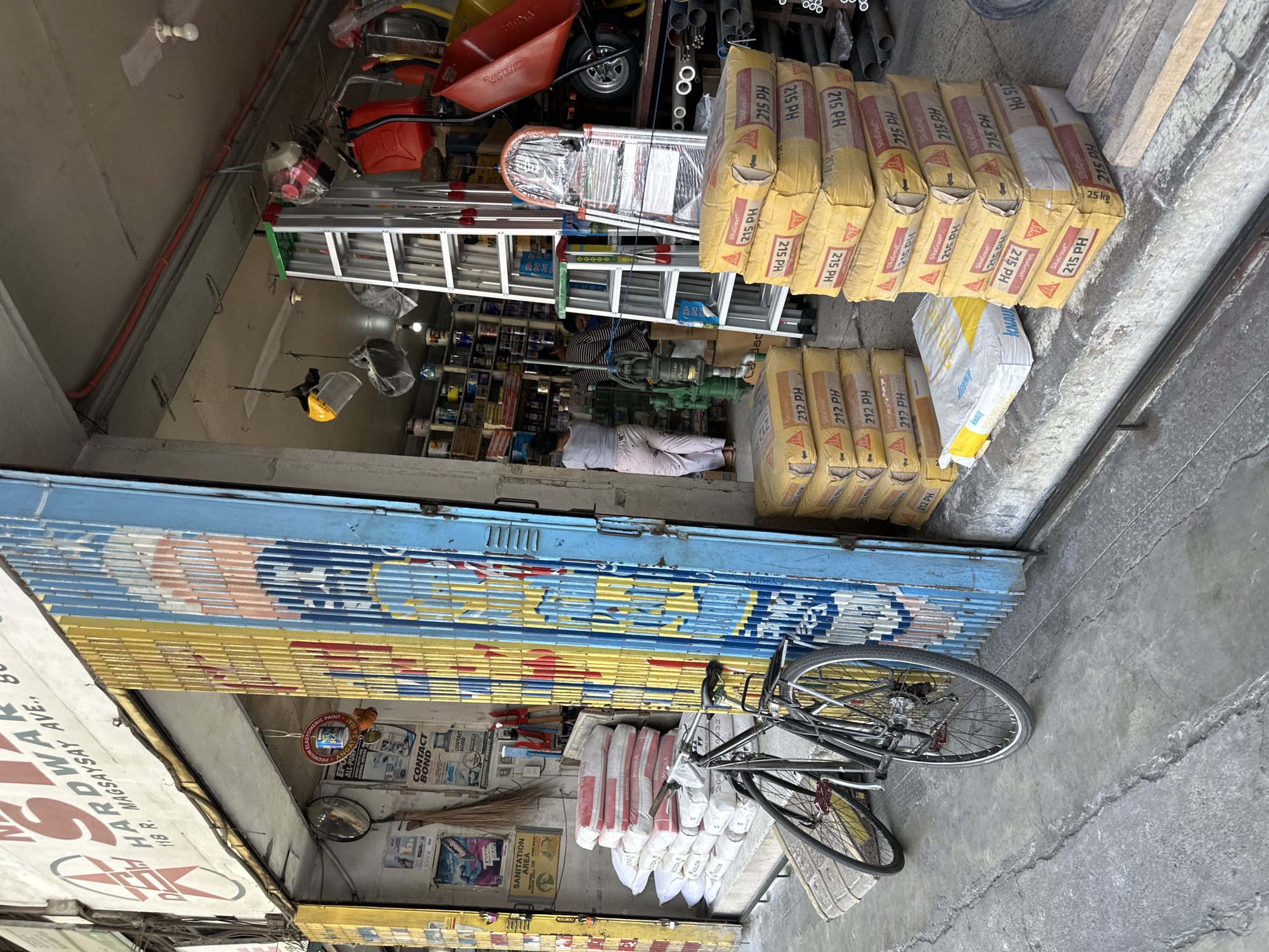 A hardware store in Davao City, Philippines, is shown in this medium shot. The exterior of the store features a blue and yellow metal gate, a sign that reads "STAR HARDWARE," and a stack of sacks of cement. A black bicycle is parked in front of the store, leaning against a stack of cement sacks. Inside the store, shelves are stocked with various hardware items, and ladders are displayed. Two women and a man are visible inside the store, with one woman appearing to be a customer and the others likely employees. The pavement in front of the store is cracked and worn, indicating a well-trafficked area. The overall scene depicts a typical neighborhood hardware store in the Philippines.