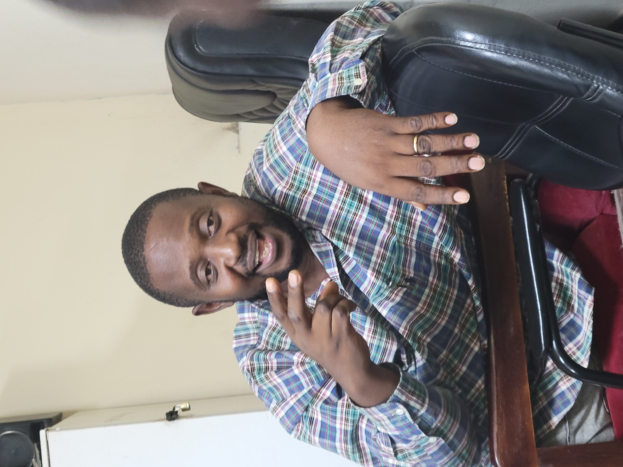 A man with dark skin and short hair is sitting on a chair. He is wearing a long-sleeved, button-down shirt with a blue, green, and purple plaid pattern. A gold ring is visible on his left ring finger. He is smiling and holding up two fingers in a peace sign with his left hand. He has a beard. The chair he is sitting on is a brown wooden chair with a black leather seat. Behind the chair is a light beige wall. The location is unknown; there is no information about Abuja, Nigeria.